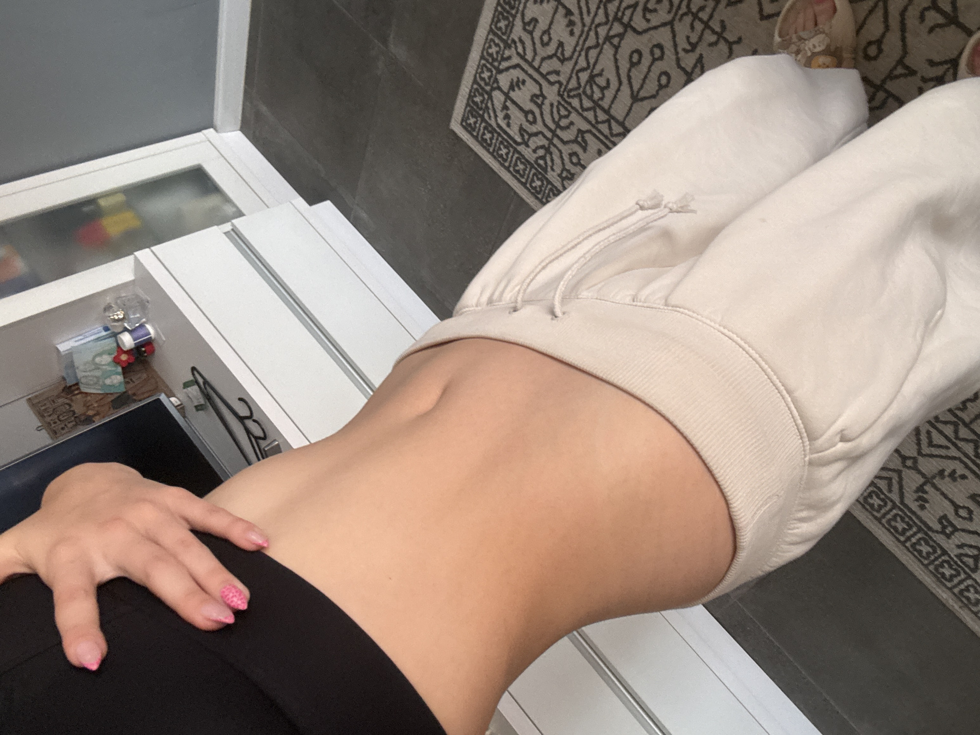 A close-up shot focuses on the midriff and lower torso of a person wearing a black crop top and light-colored sweatpants. Their hand, adorned with bright pink nail polish, rests on their stomach. The background includes a white cabinet with drawers and a glass-fronted cabinet containing various items, suggesting an indoor setting, possibly a bedroom or living area. The floor is tiled in a dark gray, with a patterned rug visible. The scene does not contain any specific landmarks or elements that would indicate a location in Sofia, Bulgaria.