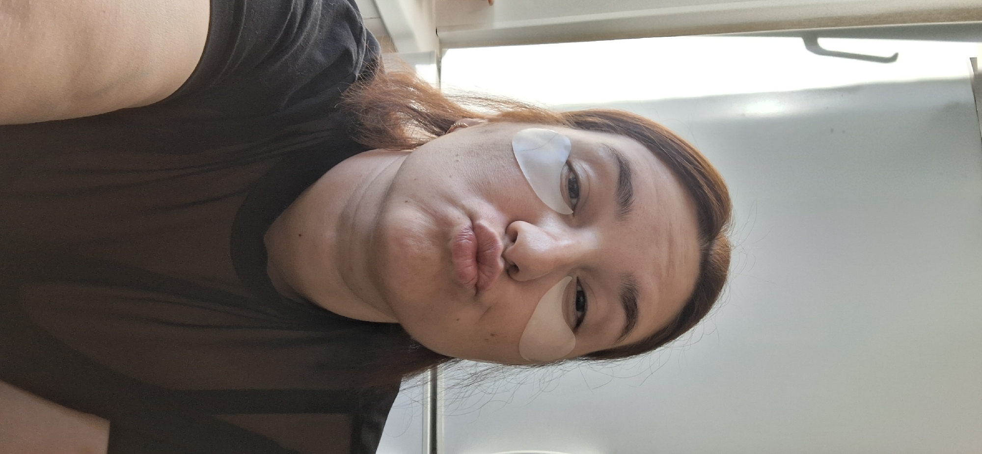 A person with reddish-brown hair and eye patches poses for a selfie with pursed lips, wearing a black, semi-sheer shirt. The background includes a white wall and window frame. The location is unknown.
