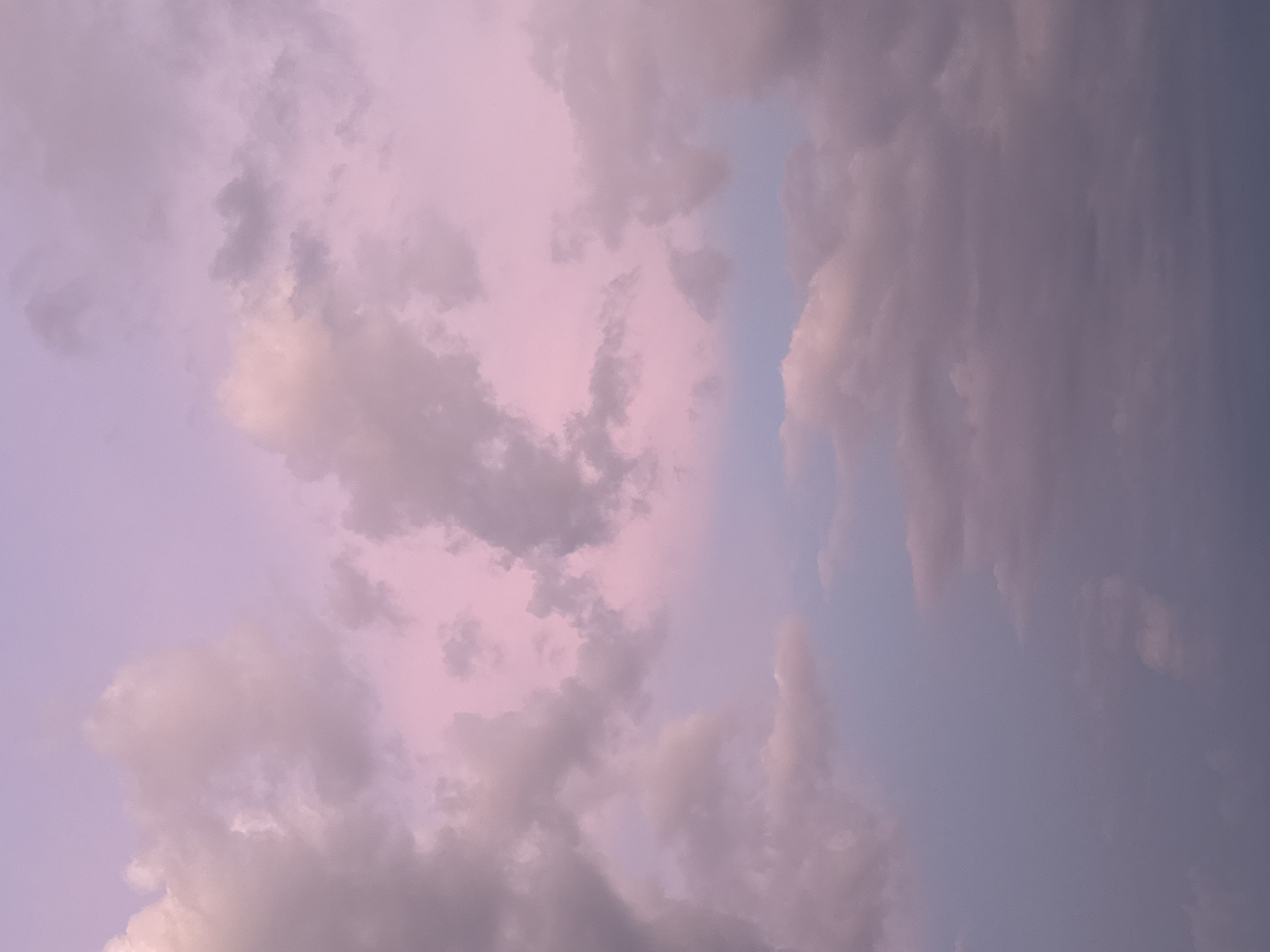 The image depicts a sky filled with clouds, likely at sunset or sunrise due to the soft, pastel hues of pink and purple. The clouds are varied in shape and density, with some appearing wispy and others more substantial and textured. A narrow band of lighter blue sky is visible between patches of cloud on the right side of the frame. The overall impression is one of tranquility and atmospheric beauty. There are no people, objects, or text visible in the image. The scene is purely a natural sky formation.