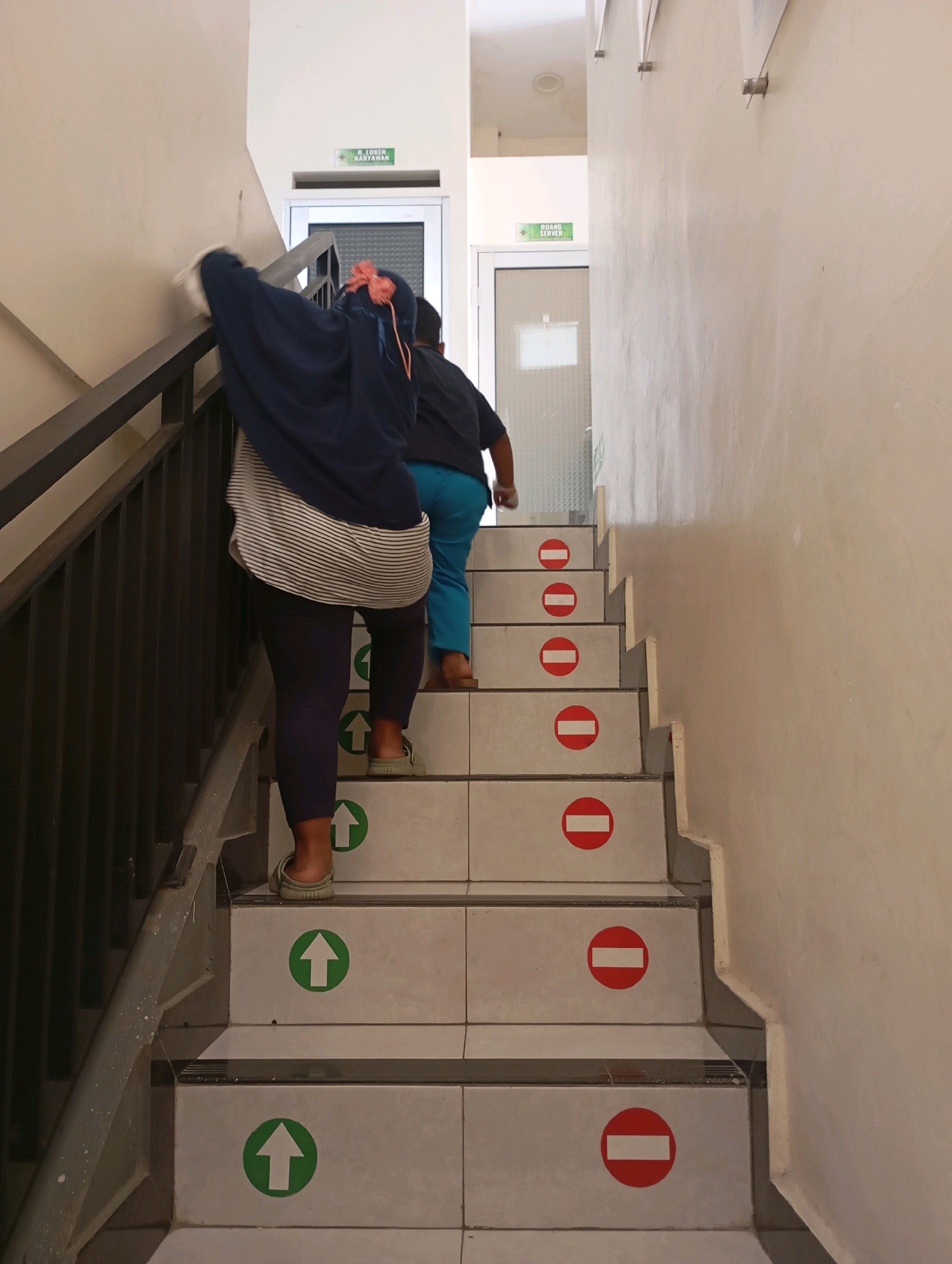 In Babelan, Indonesia, two individuals are ascending a set of indoor stairs. The person in the foreground, wearing a dark blue headscarf and a striped shirt, is on the lower steps. Behind them, a second person in a dark shirt and blue pants is on a higher step. The stairs are tiled with a white and gray pattern, and each step is marked with either a green upward-pointing arrow or a red circle with a white horizontal bar, indicating directional flow or prohibition. The stairway has a black metal railing on the left and a plain beige wall on the right. Above the stairs, doorways are visible with signage indicating "RUANG SERVER" and "R. LOKIR KARYAWAN". The lighting is ambient and appears to be from artificial sources.