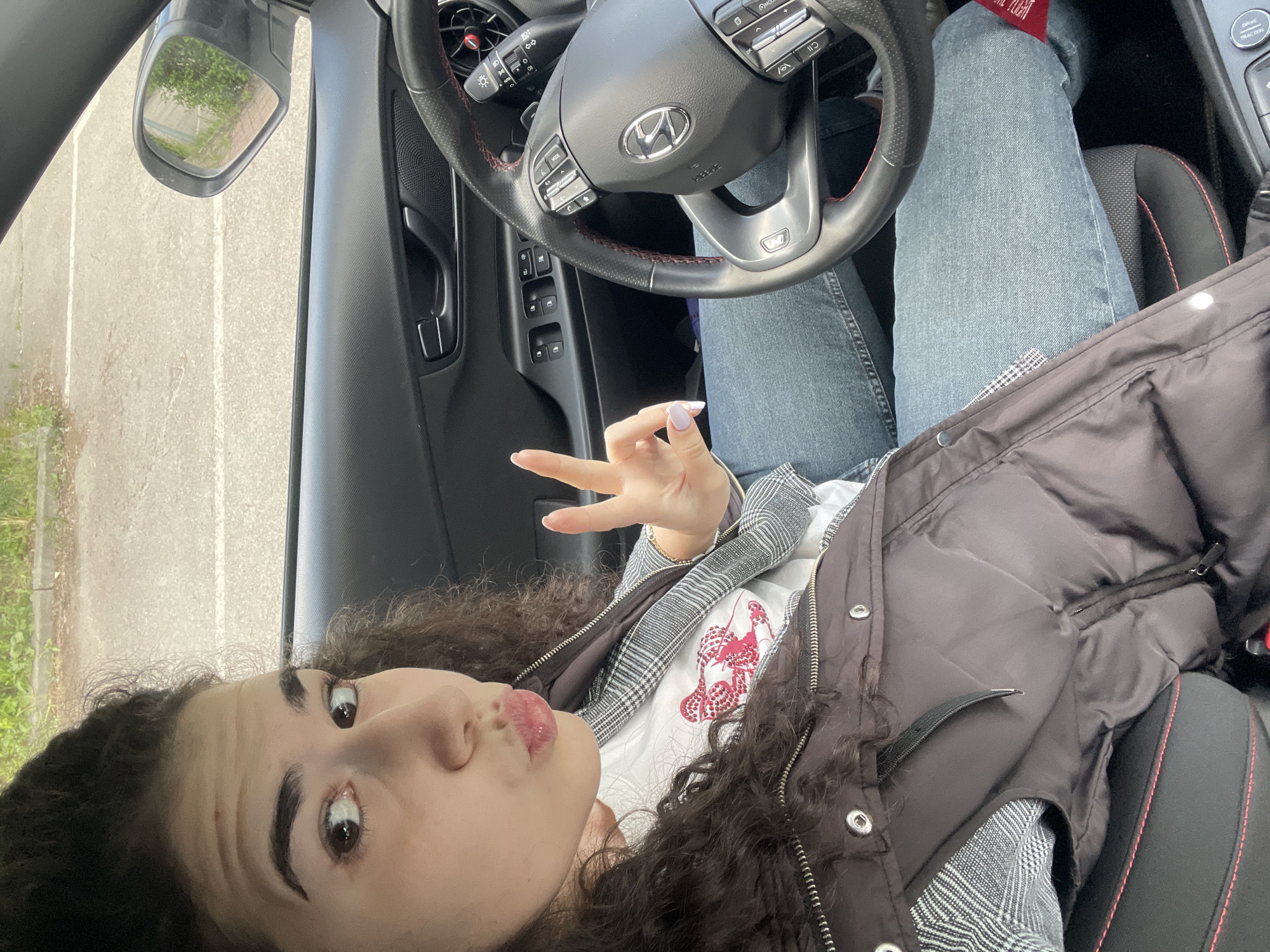 A young woman with curly dark hair is in the driver's seat of a car, making a peace sign. She is wearing a white t-shirt with a red design, a gray plaid jacket, and a black puffy vest. She is wearing blue jeans and a red sock is visible above her right leg. The car has a black interior with red stitching on the seats and steering wheel. The steering wheel has a Hyundai logo. The driver's side mirror is visible, reflecting a parking lot with green foliage in the background. The location is unidentified and cannot be determined to be Sofia, Bulgaria, based on the provided image.