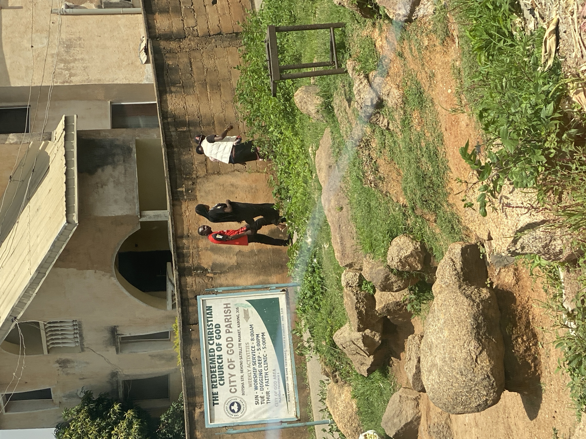 The image depicts an outdoor scene in Jos, Nigeria, focusing on a sign for The Redeemed Christian Church of God, City of God Parish. The sign, which is white with blue and green accents, is positioned in front of a rough-hewn brick wall. It lists the church's location as "BOTSHA STR, BEHIND SATELLITE MARKET, KABONG, JOS" and details weekly activities including "SUNDAY - WORSHIP SERVICE - 8:00AM", "TUESDAY - DIGGING DEEP - 5:00PM", and "THURSDAY - FAITH CLINIC - 5:00PM". Below this, it states "CITY OF GOD AREA, PLATEAU PROVINCE 1" and includes a biblical reference: "JESUS CHRIST THE SAME YESTERDAY, TODAY & FOREVER - HEB. 13:8".
In front of the wall and sign, three individuals are visible. The person on the left wears a red t-shirt and dark pants, and appears to be looking towards the right. Next to them, a person in a black garment is walking away from the camera. To the right of these two, a person wearing a white vest over dark clothing and a dark cap is walking towards the right, seemingly interacting with the person in black.
The environment is characterized by a grassy, uneven terrain with several large rocks scattered throughout. In the background, a multi-story building with a beige-colored exterior and a corrugated metal roof is partially visible, with arched architectural features and a balcony. The overall lighting suggests a bright, daytime scene. A small, rusted metal frame structure is also present in the middle ground.