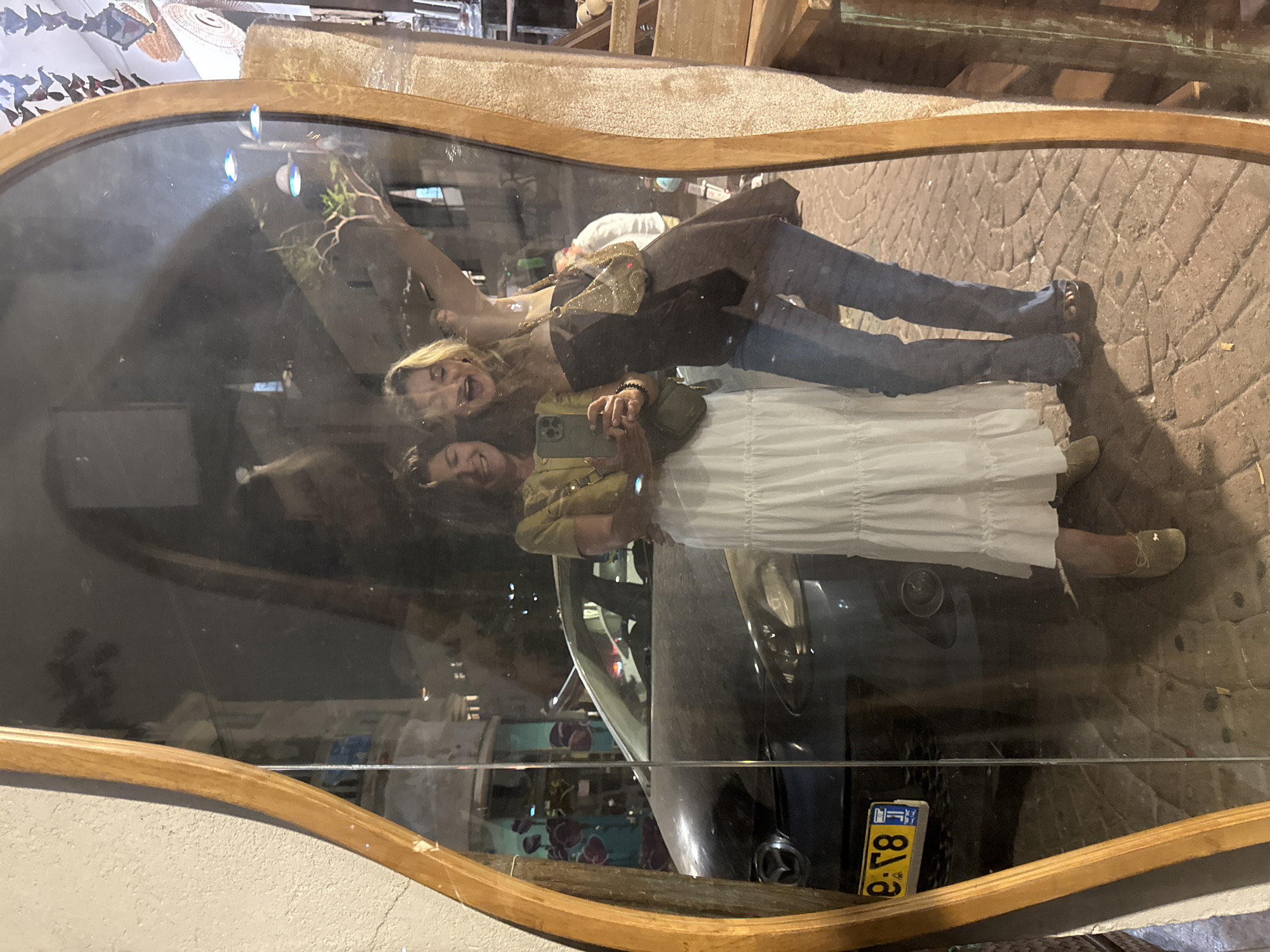 The image displays a reflection of two women captured in an irregularly shaped mirror with a wooden frame, situated outdoors at night. The scene is located on a cobblestone street in Tel-Aviv, Israel.
The woman on the left, with dark hair, wears a yellow-green short-sleeved top, a long white tiered skirt, and light-colored flat shoes. She holds a dark-colored smartphone, appearing to take the selfie, and has a dark green sling bag. She is smiling.
The woman on the right, with blonde hair, wears a dark strapless top, blue jeans, and dark open-toed sandals. She carries a small, sparkling gold handbag. Her right arm is raised, and she has an expressive, open-mouthed smile, suggesting laughter or excitement. Both women stand in close proximity.
In the background, visible through the reflection, a dark-colored Mazda car is parked on the street. Its license plate, partially visible, displays the Hebrew letter "פ" followed by ":r8" and a "JT" sticker. The street is paved with distinctive interlocking pavers or cobblestones. Reflections of distant buildings, streetlights, and a small tree are also present. To the far left, behind the parked car, a building wall exhibits abstract reddish-purple shapes on a light blue-green background, indicative of street art. To the right of the women, within the mirror's view, a textured, light-brown display stand or furniture piece is partially visible, along with hanging decorative elements.
