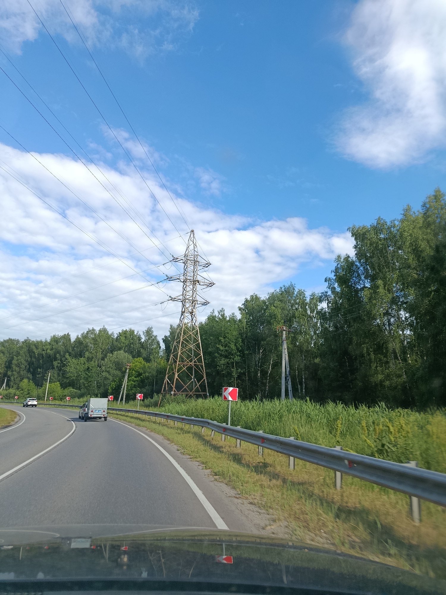 This media file captures a view from a vehicle traveling along a road in Тополиный, Russia. The scene depicts a sunny day with a blue sky dotted with white, fluffy clouds. A large, lattice-style electrical tower stands prominently on the right side of the road, with multiple power lines stretching across the sky.
The road itself is paved and curves to the left. There are two vehicles visible: a white car in the distance on the left lane and a white trailer being towed by another vehicle in the right lane. The road is bordered by a metal guardrail on the right, with tall green grass and trees forming a dense backdrop. Red and white directional signs indicating curves are visible along the roadside. The overall impression is that of a typical summer day in a rural or semi-rural area, with a focus on the infrastructure and natural landscape. The time of day appears to be late morning or early afternoon, given the bright sunlight and clear sky.