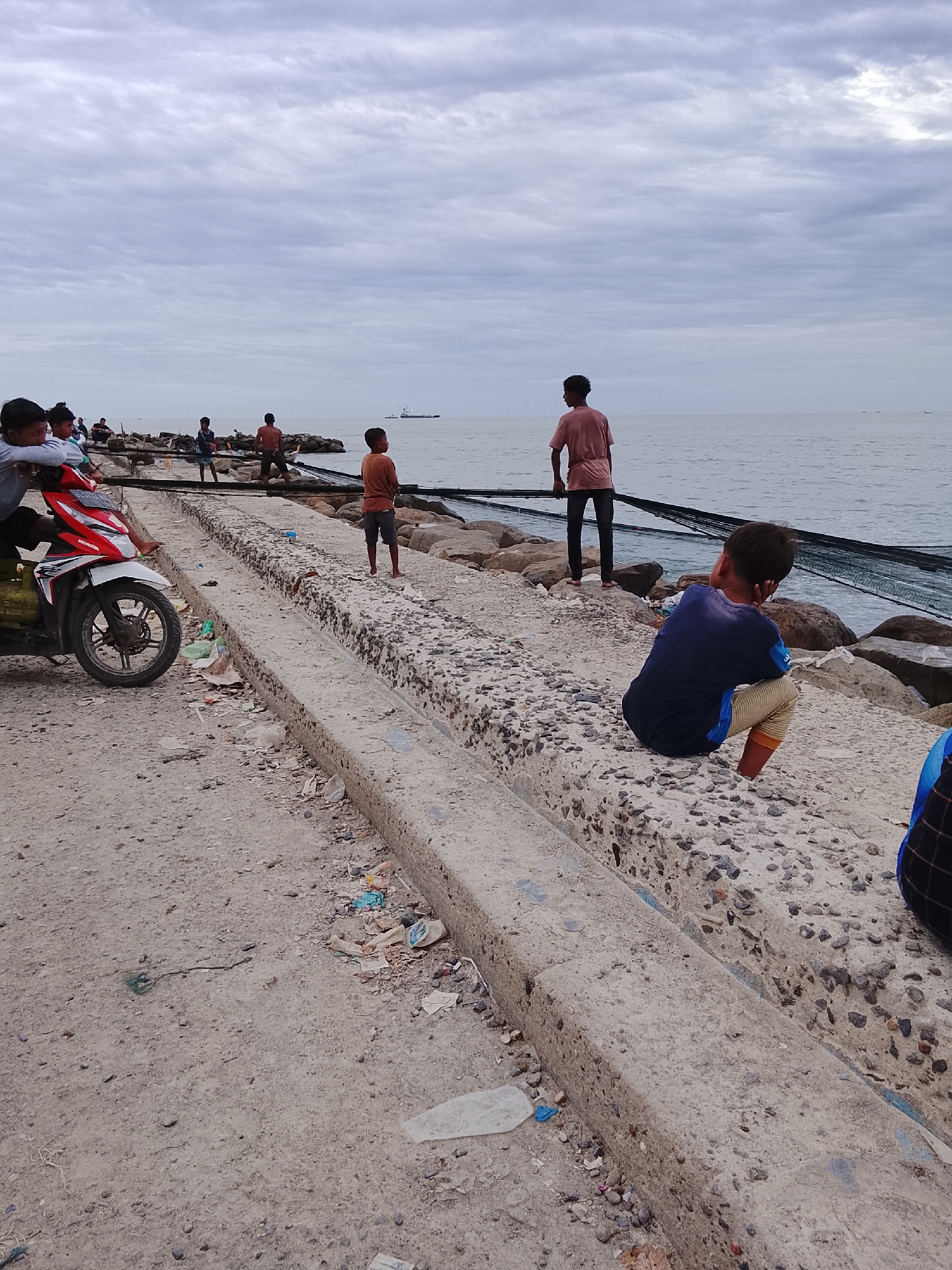 The image captures a coastal scene in Lhokseumawe, Indonesia, under a cloudy sky. Several individuals, mostly young, are present. One person sits squatting on a concrete structure while several others stand on a partially constructed seawall or breakwater made of concrete and rocks. A motorcycle is parked to the side, ridden by one person and approached by another. There is visible litter scattered on the ground. In the background, a dark net structure stretches along the rocks in the ocean. A cargo ship can be seen in the distance.