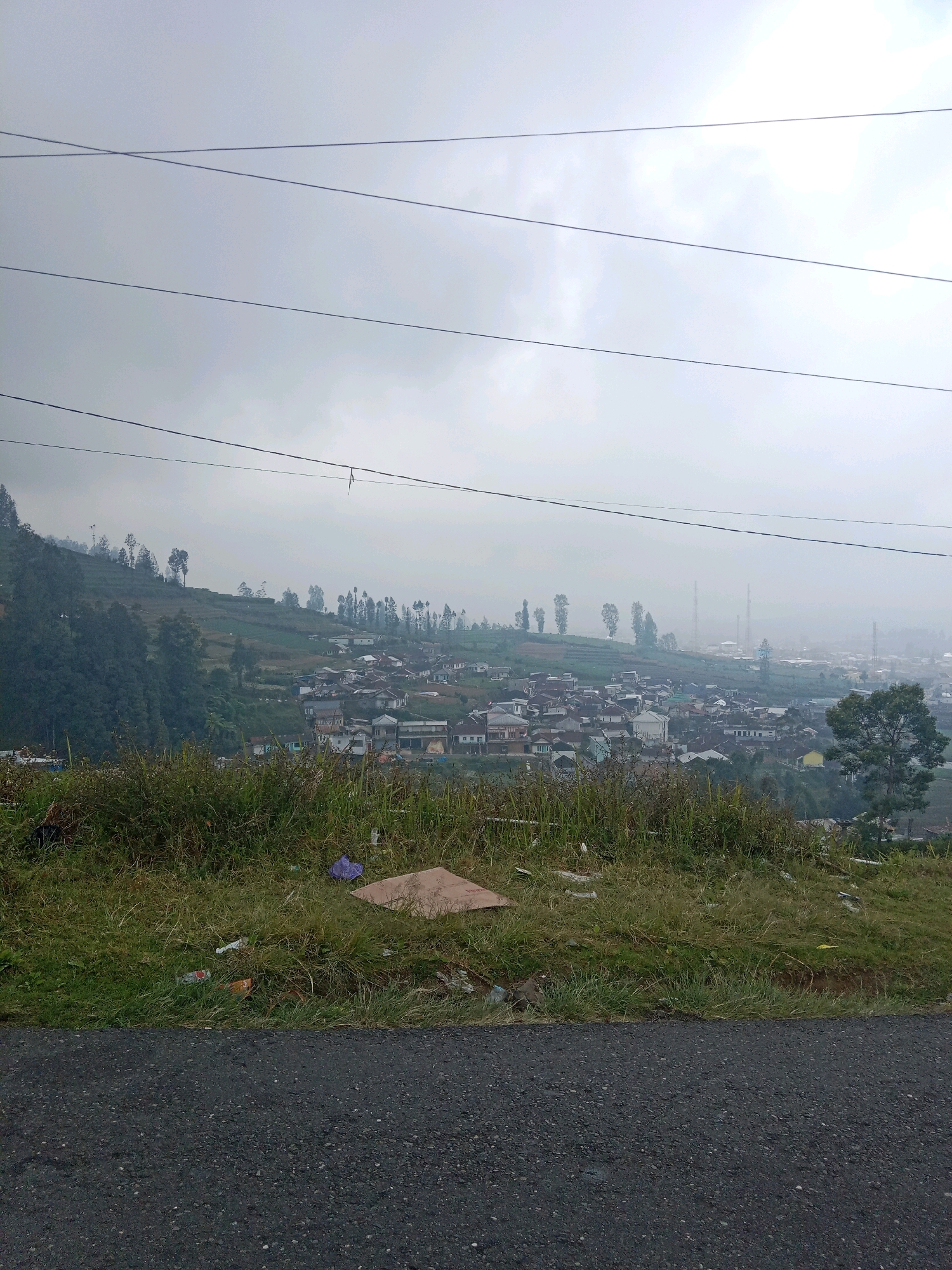 The image shows a view of a village nestled in a valley in Batur, Indonesia. The sky is overcast, and a thin layer of haze obscures the distant landscape. Power lines are visible crisscrossing the upper portion of the frame. In the foreground, a patch of tall, dry grass and weeds is littered with various pieces of trash, including plastic bags, paper, and cardboard. A paved road occupies the bottom of the image. The village itself consists of numerous small buildings with varied roof colors, clustered together on the slopes of the surrounding hills. The hills are covered with a mix of trees and agricultural terraces. In the far distance, industrial structures and more hazy atmospheric conditions are visible. There are no people or animals clearly visible in the scene.