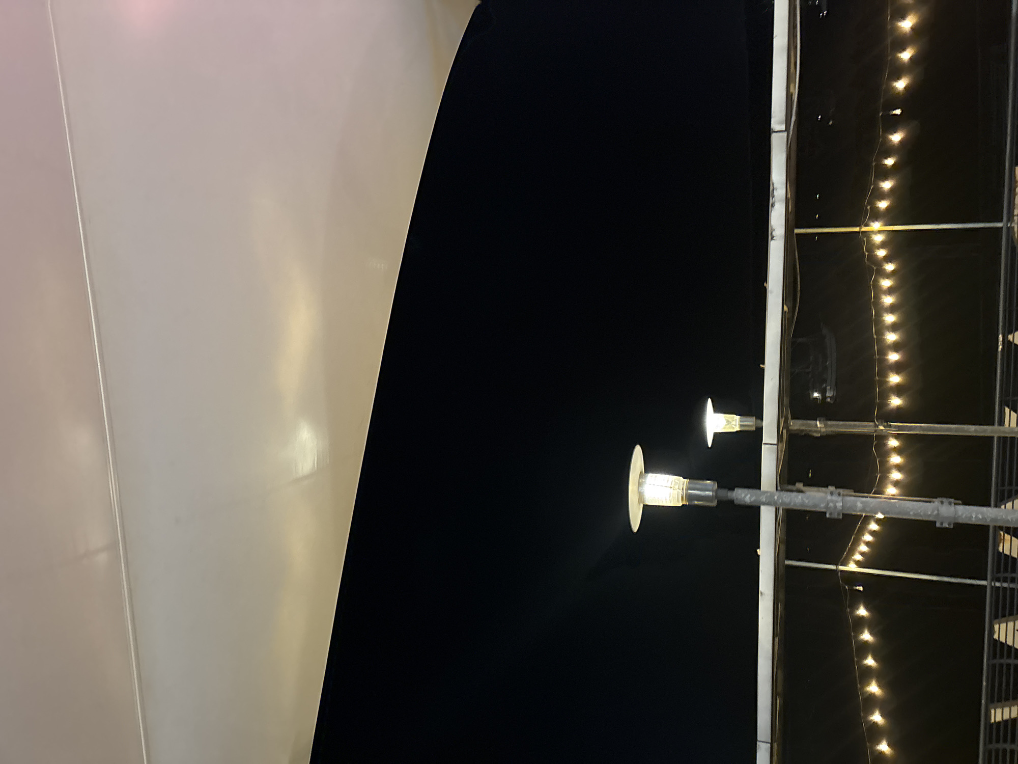 The image depicts an outdoor scene at night, likely at a waterfront establishment in North Jakarta, Indonesia. A large, curved canopy or awning, appears to be made of a light-colored, possibly translucent material, dominates the upper portion of the frame, casting a soft glow. Below the canopy, the sky is a deep, uniform black, indicating nighttime.
In the mid-ground, two modern-style lampposts are visible. The lamppost on the left is closer and more prominent, with a cylindrical, textured shade housing a bright, warm light. This lamppost is mounted on a metal pole with visible fixtures. A second, identical lamppost is positioned further to the right and slightly behind the first, also illuminated.
Below the lampposts, a horizontal barrier or railing is present, with a dark, reflective surface, possibly water or a dark-colored surface. Strung along this barrier are numerous small, warm-toned lights, resembling fairy lights or string lights, creating a festive and inviting ambiance. These lights are spaced evenly and appear to be slightly out of focus, contributing to the nighttime mood.
In the foreground, a portion of a railing or structure is visible at the bottom, made of dark metal bars, suggesting the viewer is on a raised platform or walkway. The overall impression is of a serene, possibly dining or recreational area illuminated by artificial light against the dark night sky.