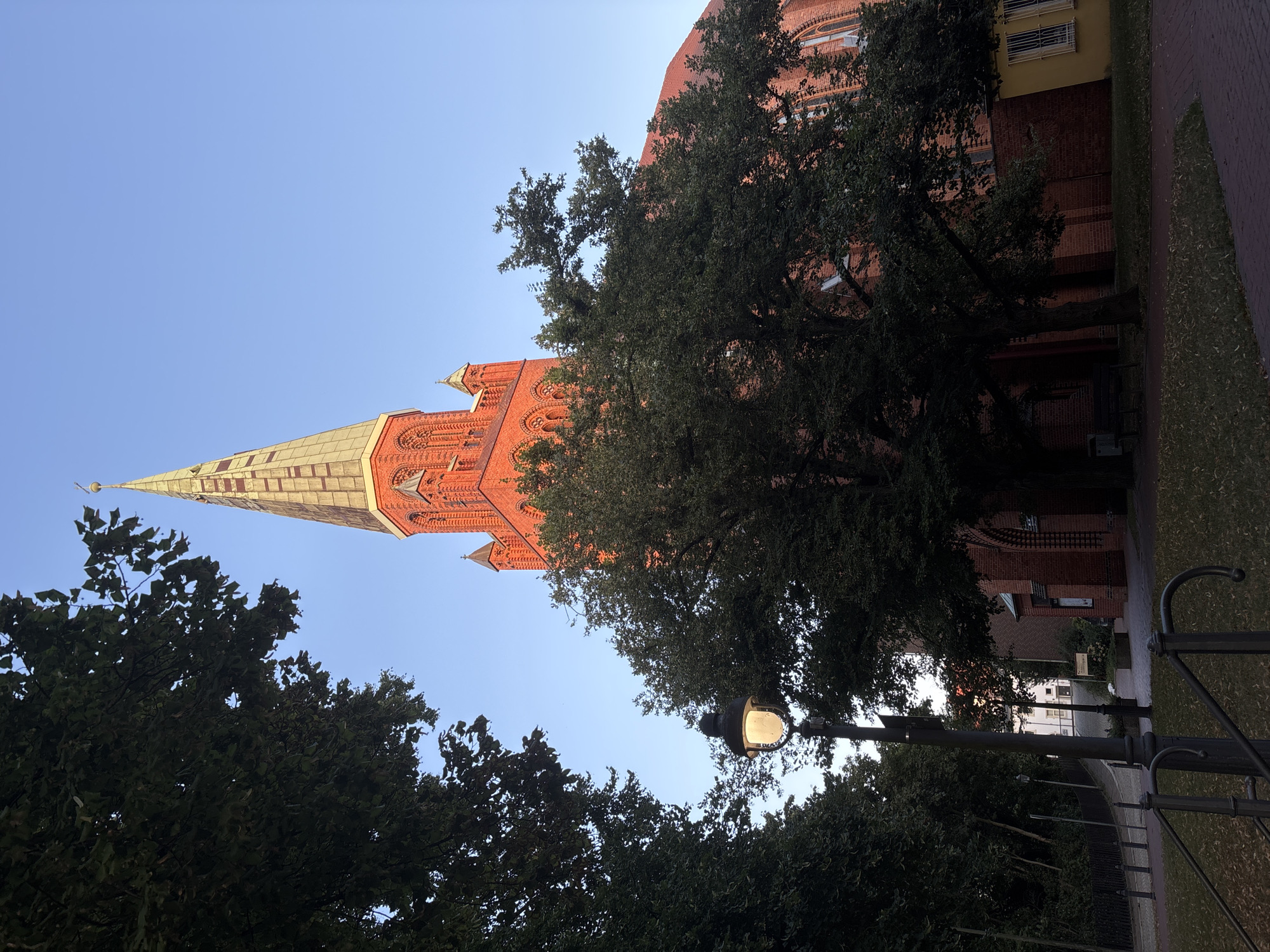 This is a medium shot, outdoor, vertical photograph taken during daylight. The scene depicts a brick church with a tall, pointed steeple that is partially obscured by large trees in the foreground. The steeple is capped with a golden-colored spire. To the left of the steeple, a streetlamp with a glowing bulb is visible. The surrounding environment consists of a paved walkway and grassy areas. The sky is a clear, light blue. The overall impression is that of a serene urban setting featuring a prominent architectural landmark.