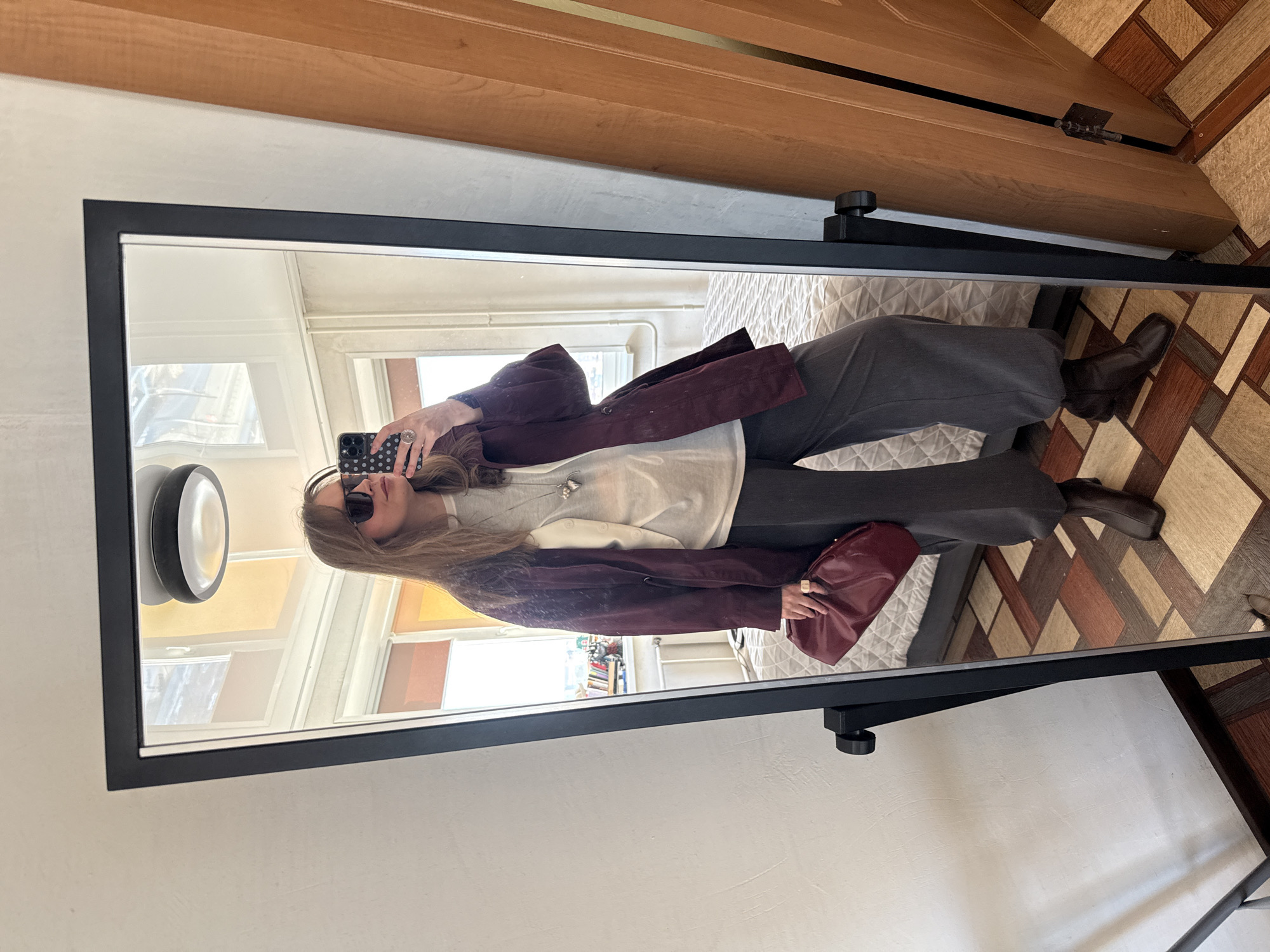 A person is reflected in a standing mirror. The person is wearing sunglasses, a light-colored sweater with a necklace, dark trousers, and brown boots. They are holding a maroon purse and a cell phone. The mirror's reflection shows a room with a window, books, and white walls. The room is lit by natural light. The floor around the mirror is patterned. The scene is indoors. There is no indication of the location being Izhevsk, Russia.