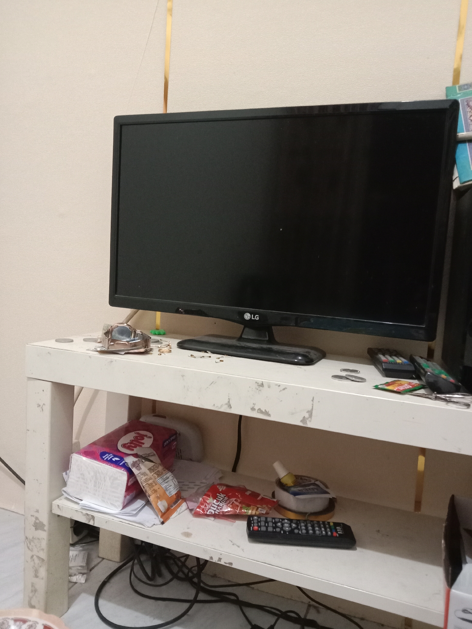 This image depicts a white metal shelf unit with a black LG monitor on top. The shelf unit is positioned against a light-colored wall with vertical gold accents. On the top shelf, to the left of the monitor, is a small, metallic object resembling a watch. There are also a few coins and some small green objects on this shelf. To the right of the monitor, on the same shelf, are two remote controls and a small package with colorful writing.

The lower shelf of the unit contains a stack of white paper products, possibly tissues, with a pink and white packaging labeled "Jelly." Next to this are two snack packages, one orange and the other red with white text. A small container with a yellow rim and a blue and white interior sits to the right, holding a small tube or bottle. A black remote control is placed in front of this container. The lower shelf also has visible black electrical cords and cables.

The overall environment appears to be an indoor setting, likely a room, given the presence of a shelf unit and electronic devices. The lighting is moderate, casting some shadows. The image is a still photograph taken at a medium distance, capturing the arrangement of objects on and around the shelf.