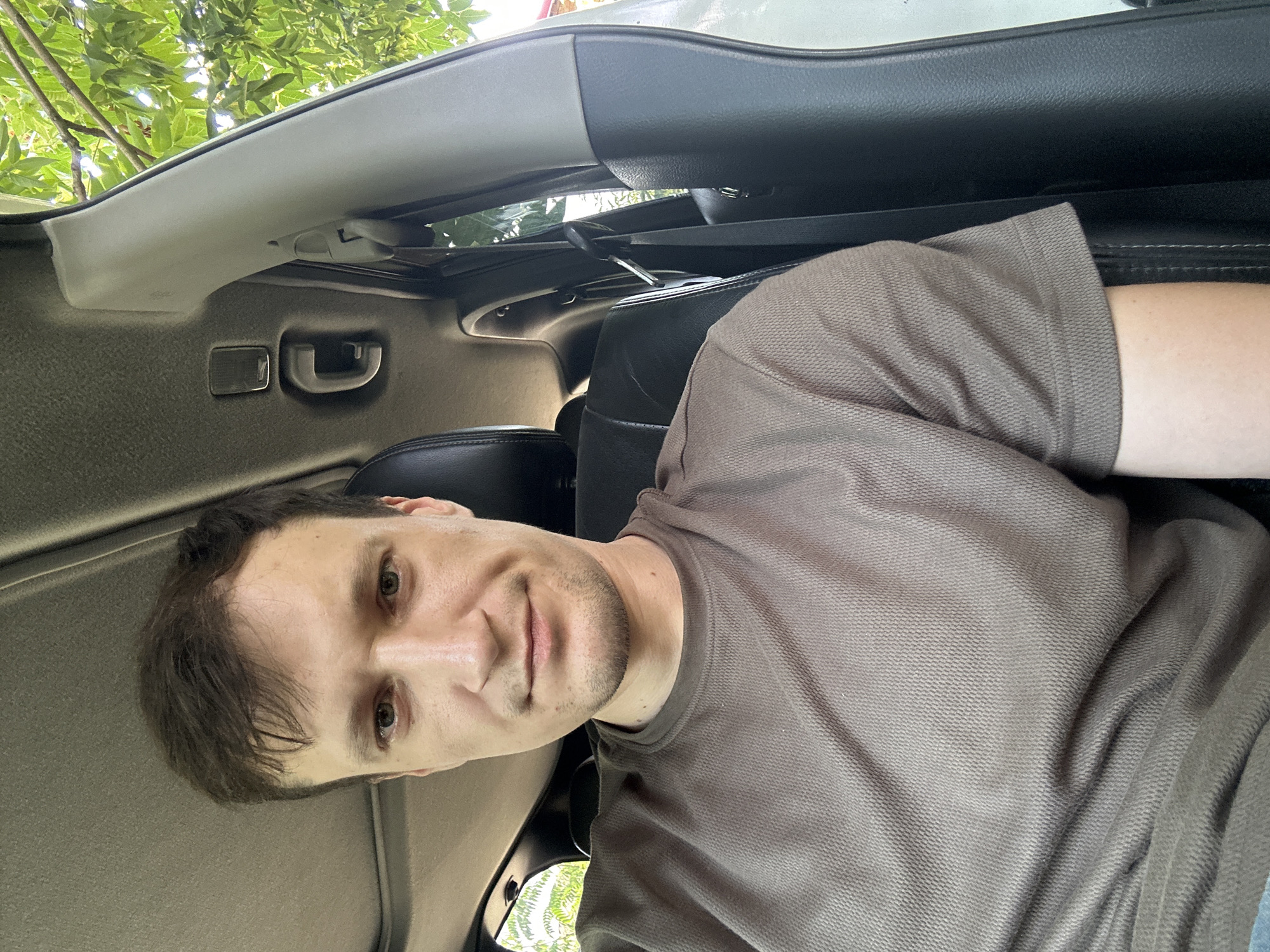 A man with short brown hair and light skin is sitting in the driver's seat of a car. He is wearing a brown t-shirt. The interior of the car is visible, including a black leather seat, a sunroof, and the car's pillar. Outside the car, green leaves from trees are visible. The image appears to be taken in the city of Makiv, Ukraine.