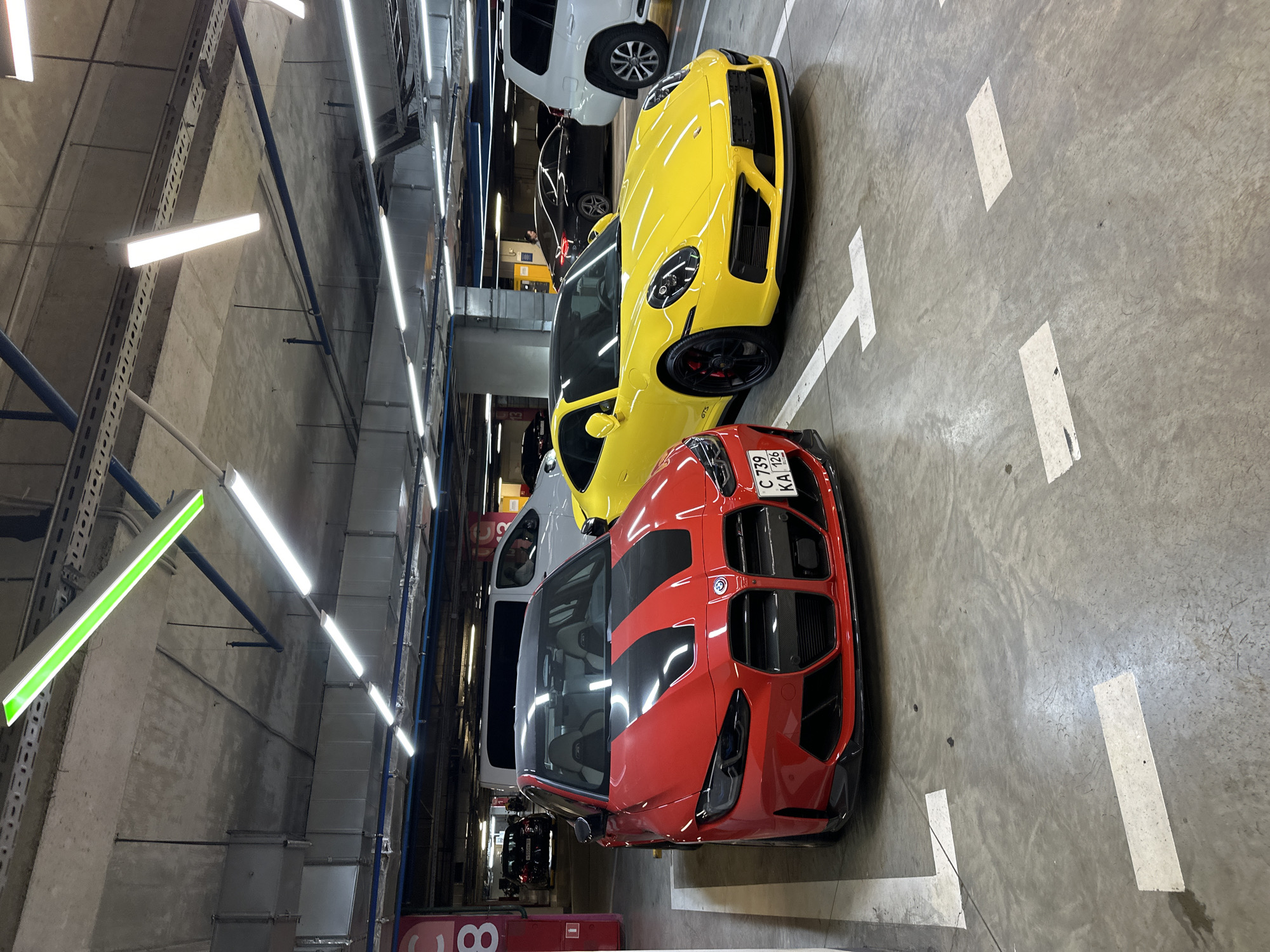 The image depicts a vibrant scene within a well-lit, modern indoor parking garage, likely in Sochi, Russia, given the license plate information. The perspective is slightly elevated, looking down at two prominent sports cars parked side-by-side, with other vehicles visible in the background.
In the immediate foreground on the right, a striking red BMW M3 or M4 (G80/G82 generation) is parked, featuring aggressive black racing stripes on its hood, a large distinctive grille, and carbon fiber accents. Its Russian license plate, "C 739 KA 193," is clearly visible, confirming its origin from Krasnodar Krai, the region of Sochi.
Adjacent to the red BMW, a bright yellow Porsche 911, possibly a GT3 model indicated by a partially visible badge on the side, is parked. It showcases black multi-spoke wheels and black exterior trim.
Behind the yellow Porsche, a white SUV, possibly a Toyota Land Cruiser, is parked, and further back, parts of other dark-colored vehicles can be seen. The parking garage floor is concrete, marked with crisp white lines delineating the parking spaces.
The ceiling of the garage features numerous bright, linear fluorescent light fixtures, providing strong illumination. Some sections of the ceiling also reveal blue pipes, metal ventilation ducts, and exposed concrete structures. Notably, one of the light fixtures in the upper left corner emits a distinct green glow, possibly indicating an exit or a special zone within the garage. A red sign with the number "8" is partially visible in the bottom left, likely indicating a parking level or section.