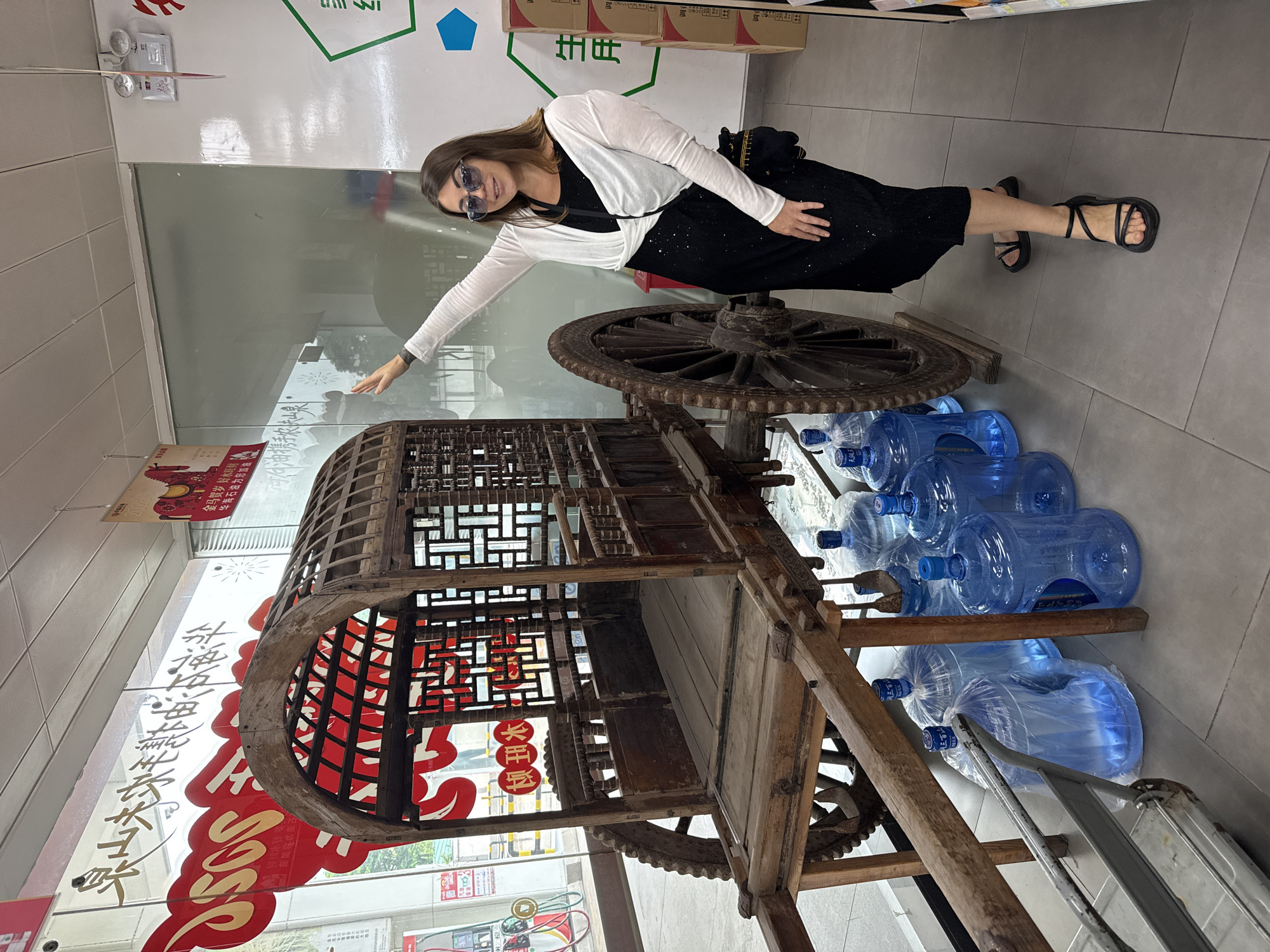 A woman stands next to a wooden ox cart with large wheels, indoors, possibly in a store or exhibition space in Huizhou, China, given the presence of Chinese characters on signs. The ox cart is old and ornately carved, with a covered seating area. It is surrounded by several large, clear plastic water jugs. The woman is wearing sunglasses, a black dress, and a white shrug. She is smiling and pointing at something above and to the left of her. The background includes shop displays and signage, one of which features a red horse graphic and Chinese characters.