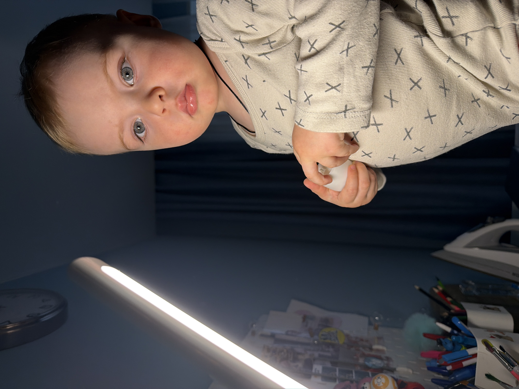 A rotated, medium shot presents a child wearing a patterned shirt, holding a white object, possibly a ball. The child has light skin, blonde hair, and light eyes. In the background are a table lamp, a clock, and various stationery items including pens and pencils, as well as an iron and other indistinct objects. The scene is indoors, and the image is dimly lit. There is no information to conclude that this image was taken in the city of Votkinsk, Russia.