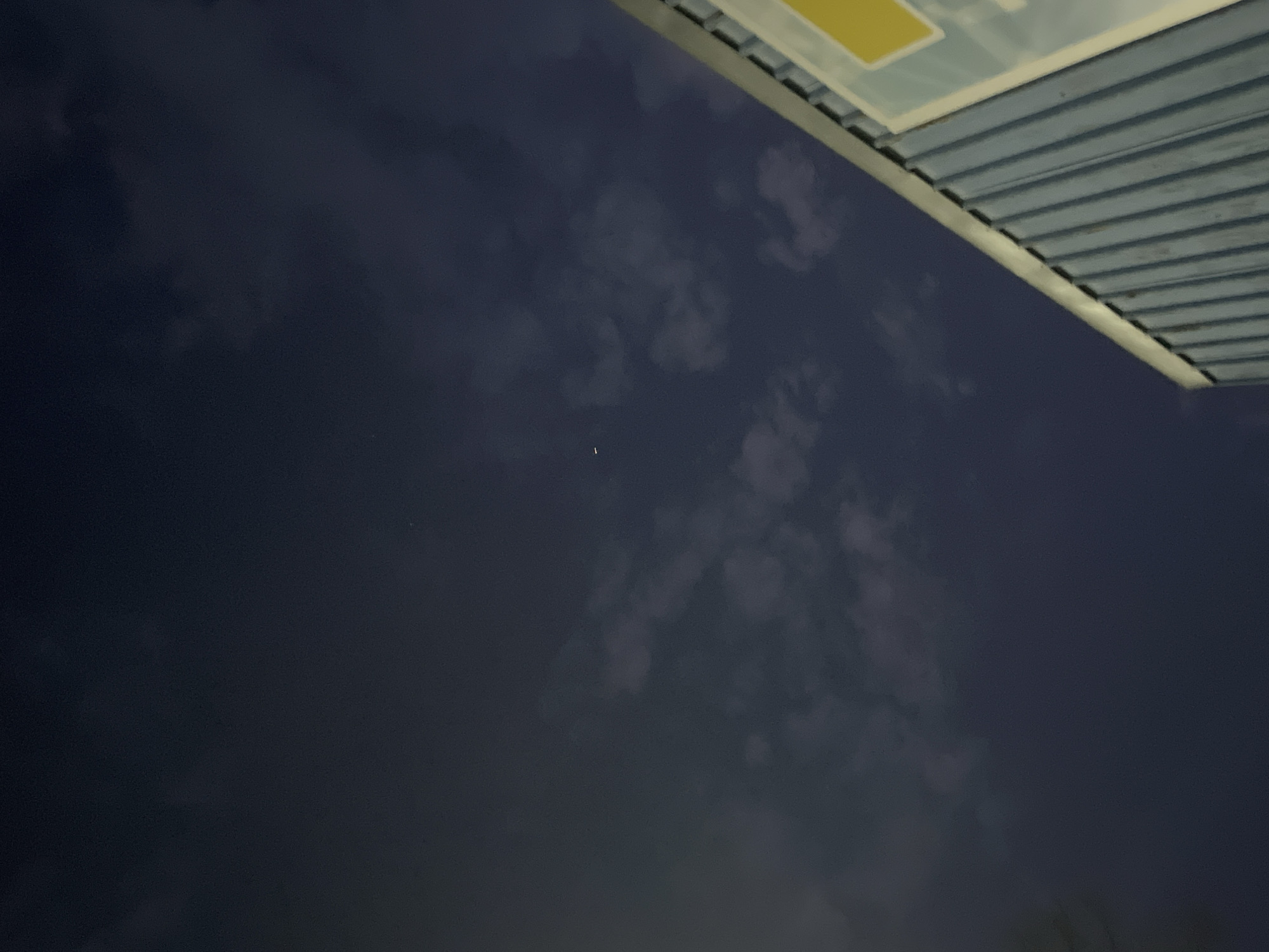 The image depicts a dark, seemingly nighttime sky. A structure with a corrugated surface, possibly a roof, is visible in the upper right corner. Faint, amorphous cloud formations are scattered throughout the sky. A single, tiny point of light, potentially a star, is observable in the center of the frame. The specific location in Elhovo, Bulgaria cannot be determined from the visual information.