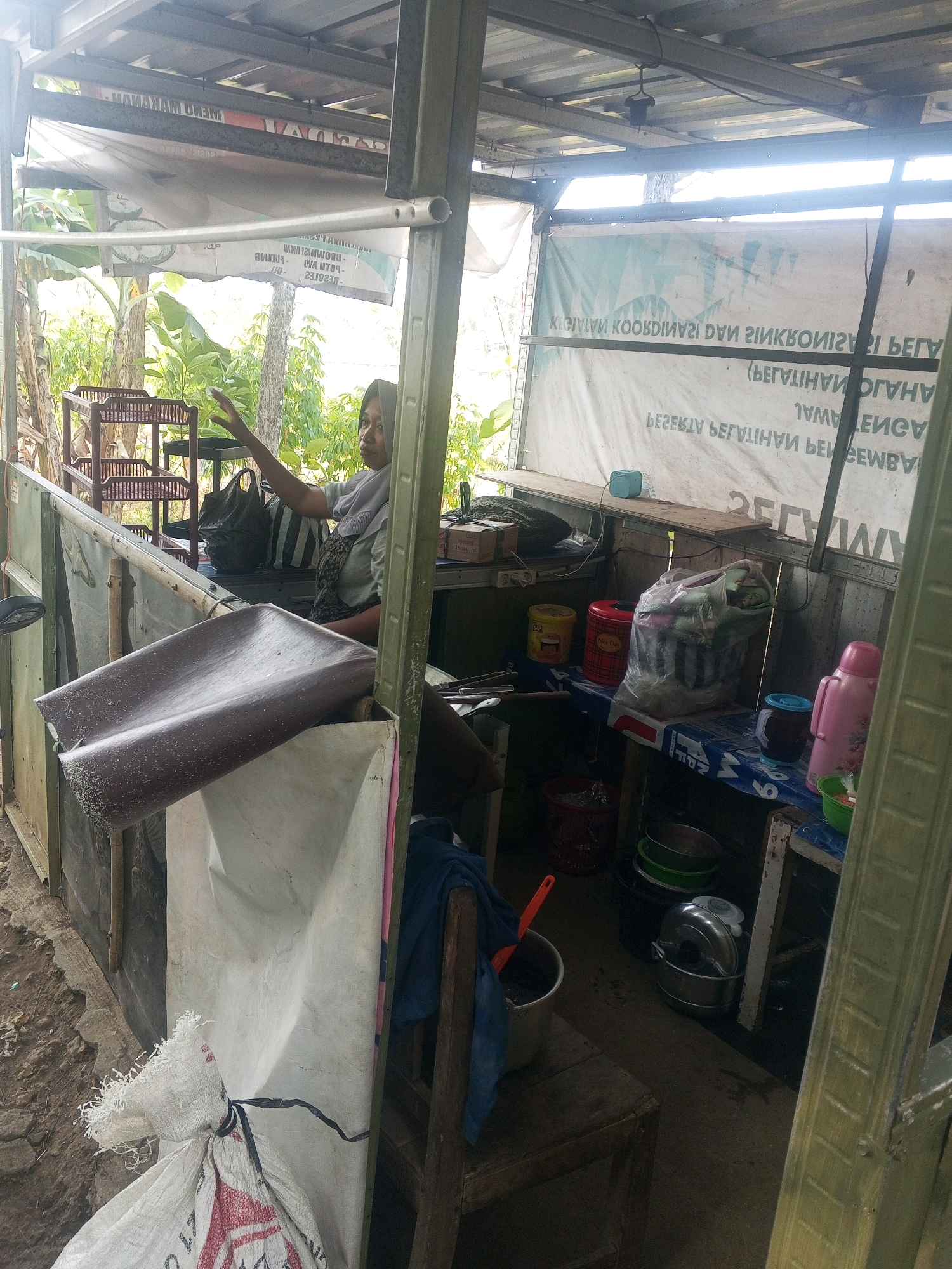 A woman wearing a hijab is visible in the background of an outdoor stall, possibly in the city of Sari, Indonesia. She is seated and gesturing with her right hand. The stall is made of metal and wood, with a corrugated metal roof. Various items are stored within the stall, including plastic bags, containers, and what appears to be a tarp. In the foreground, a white sack with red lettering is tied with black rope to a vertical support. The ground outside the stall is dirt and gravel. The lighting suggests it is daytime.