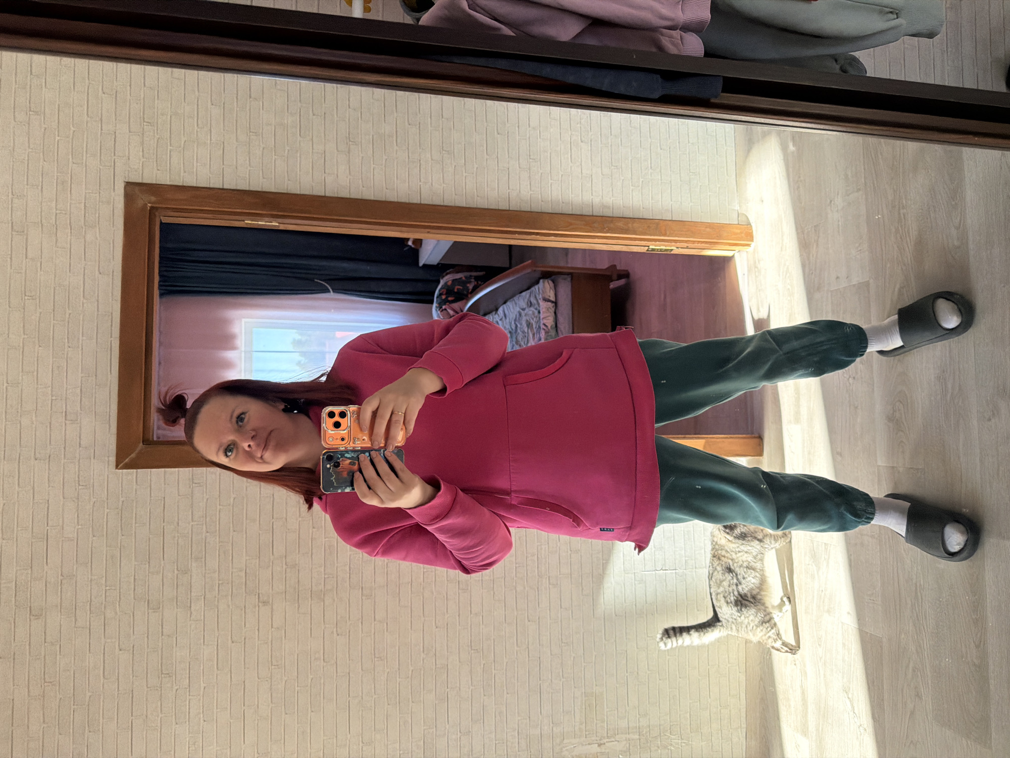 A woman is taking a selfie in a mirror. The mirror is attached to a closet door. The woman is wearing a pink hoodie and green sweatpants, along with black slides. She is holding two cellphones. In the mirror's reflection, a glimpse of a room is visible with a bed and dark curtains. The background features a wall with a brick pattern. A cat is present near the woman's feet. The photo was taken in Chaikovsky, Russia.