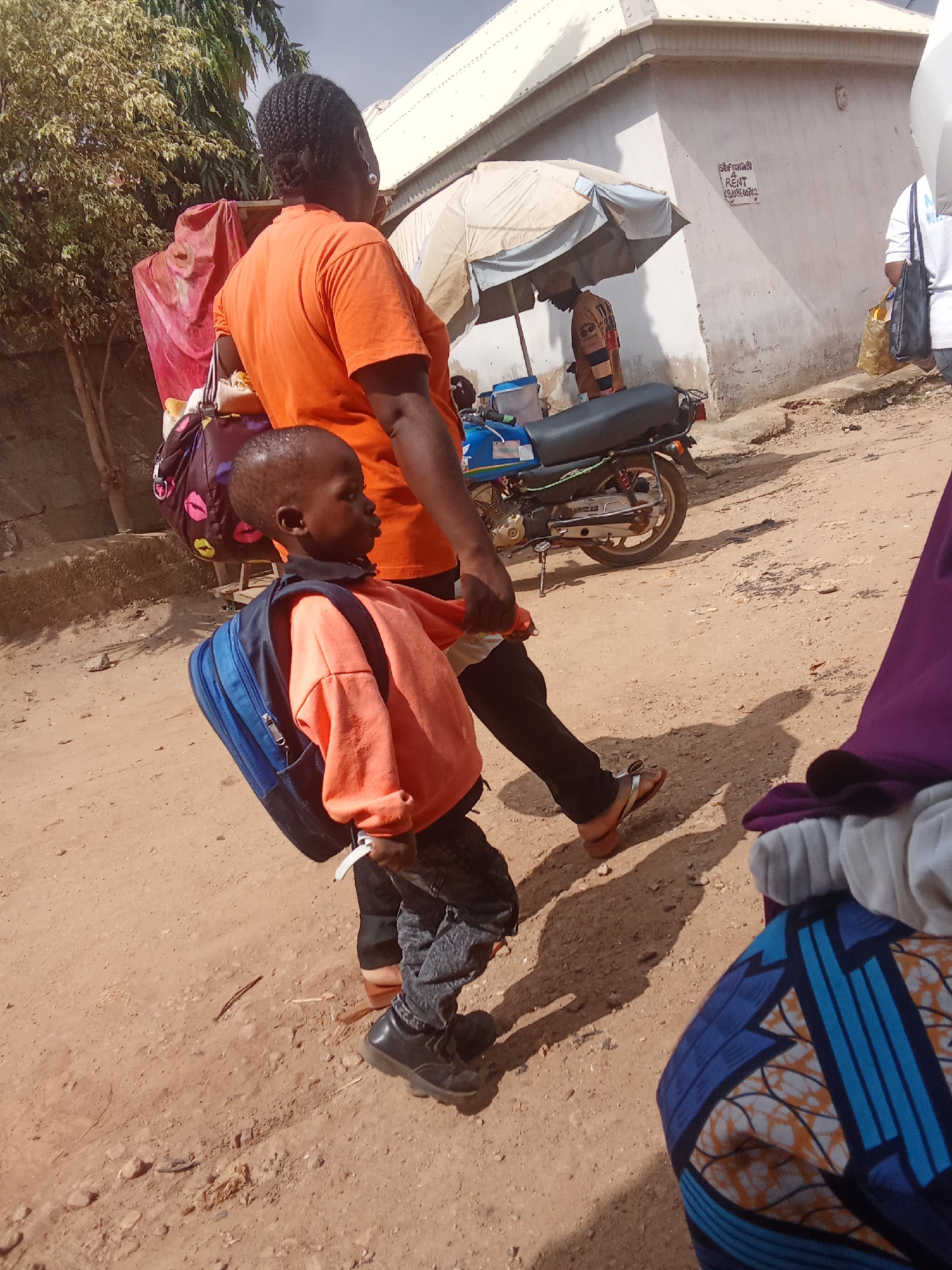 In Kagini, Nigeria, a woman in an orange t-shirt walks with a young child who is carrying a blue backpack. The woman has braided hair and is wearing sandals. The child is wearing an orange long-sleeved shirt, jeans, and black shoes. They are walking on a dirt road in front of a building. A motorcycle is parked nearby, and an umbrella shades a vendor. The scene appears to be a street in a residential or commercial area.
