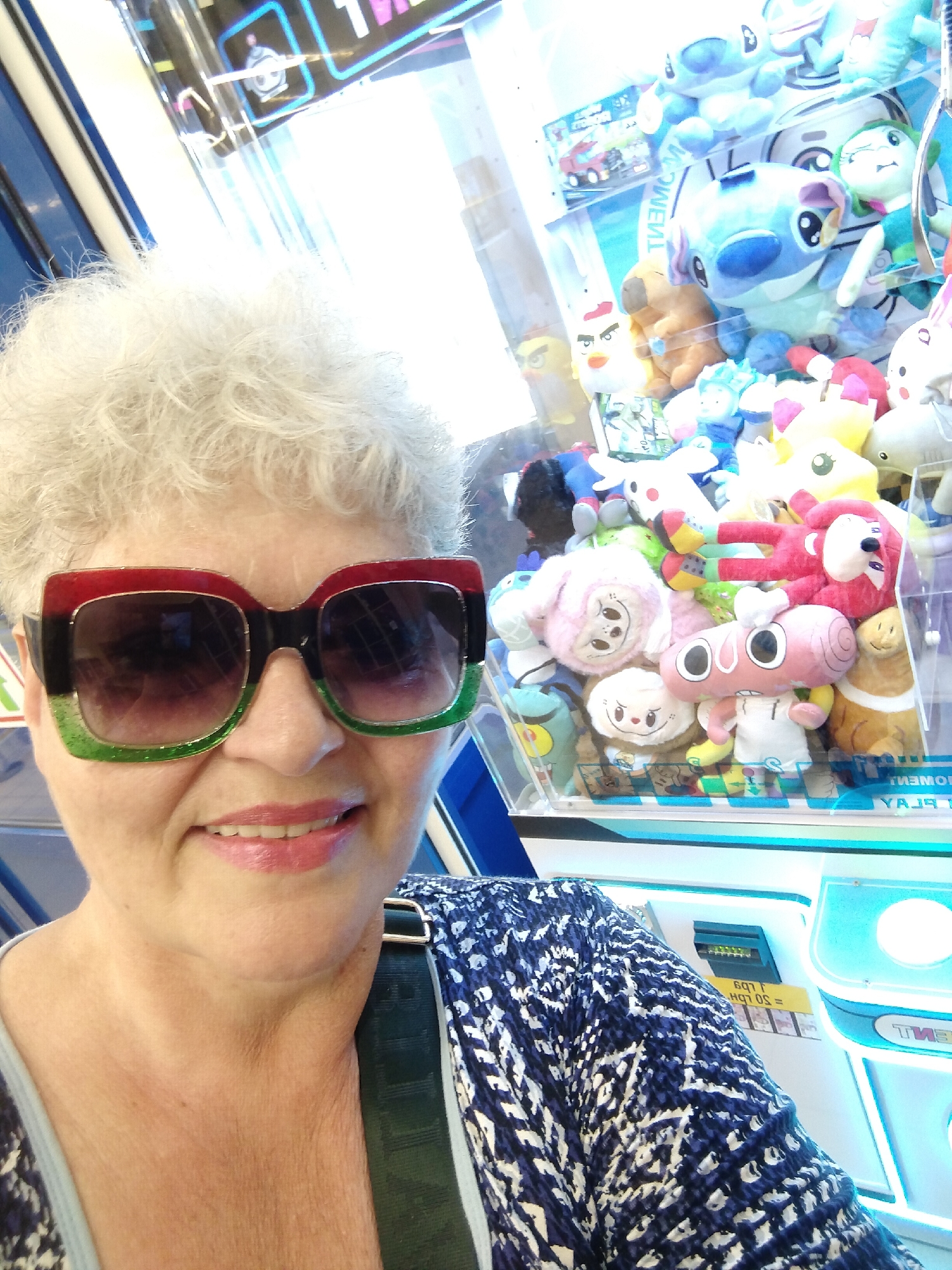 A middle-aged woman with white, curly hair and large, square sunglasses with red, black, and green frames poses for a selfie. She is smiling and has on lipstick. She is wearing a dark blue and white patterned top with a black strap visible. Behind her, a claw machine is filled with various plush toys, including Stitch from Lilo & Stitch, a tan bear, and other cartoonish figures. The machine has blue and white accents and appears to be in an entertainment or arcade setting. The setting is indoors, and the lighting suggests a bright environment. The image was taken in Horishni Plavni, Ukraine.