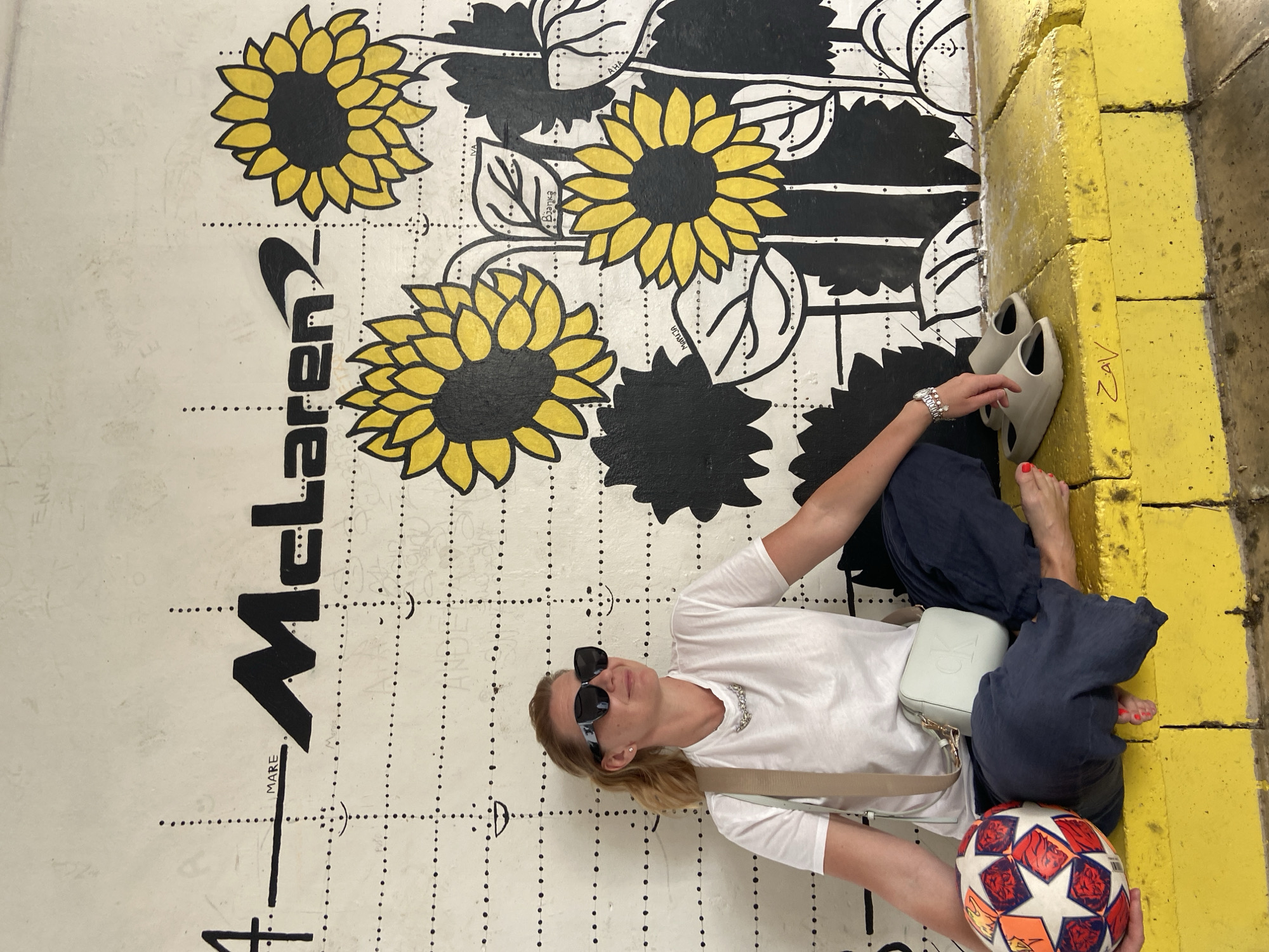 A woman is lying on the ground next to a wall in Budva, Montenegro. The wall is painted with sunflowers, black foliage, and the name "McLaren." The ground has a grid of dotted lines and a height chart. The woman is wearing sunglasses, a white t-shirt, dark blue pants, a silver necklace, and a Calvin Klein purse. She is holding a soccer ball. Beside her are a pair of open-back sandals on a yellow wall, and the graffiti tag "ZAV" is on the yellow wall.