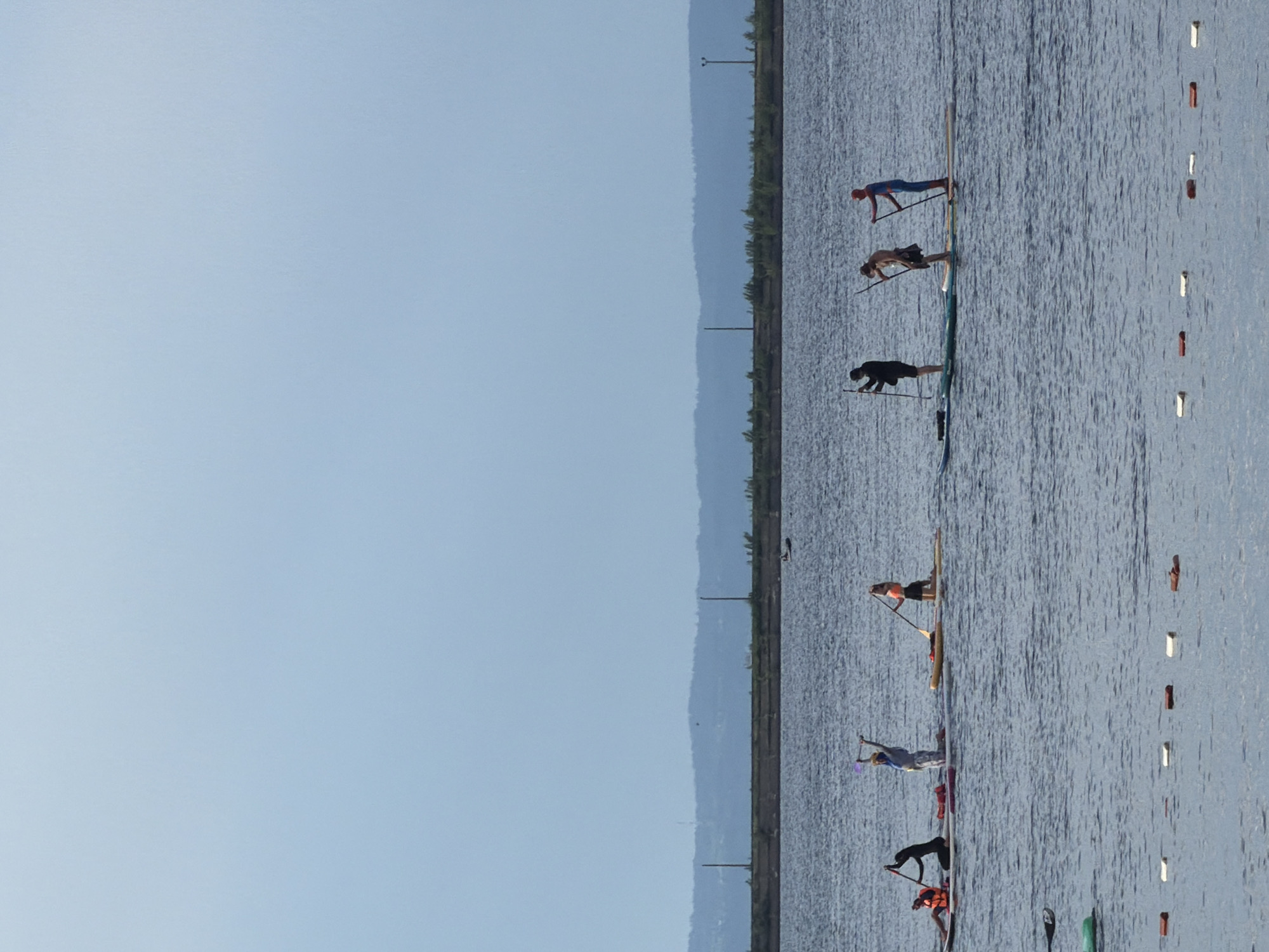 The image shows a group of people engaged in stand-up paddleboarding on a body of water. Several individuals are positioned on their paddleboards, some in a kneeling or standing posture, and one is in a plank position. The water is calm and appears to be a lake or a wide river. In the background, a shoreline with trees and some man-made structures, possibly piers or buoys, are visible. The sky is clear and light blue, suggesting a daytime setting with fair weather. The overall scene depicts a recreational activity or possibly a competition. The orientation of the image is rotated 90 degrees clockwise. The location context provided is Chaikovsky, Russia.