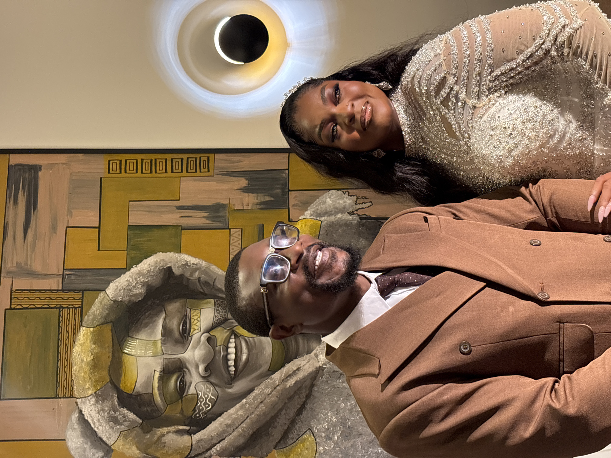 In Bar Jirgi Summa, Nigeria, a couple poses for a photo in front of a large, colorful painting. The man, wearing a brown suit, white shirt, and dotted tie, is on the left, his glasses reflecting the light. The woman, to his right, is adorned in a dazzling, beaded gown and a tiara. Behind them, the abstract painting features a portrait with tribal markings. To the right, a modern wall lamp illuminates the scene.