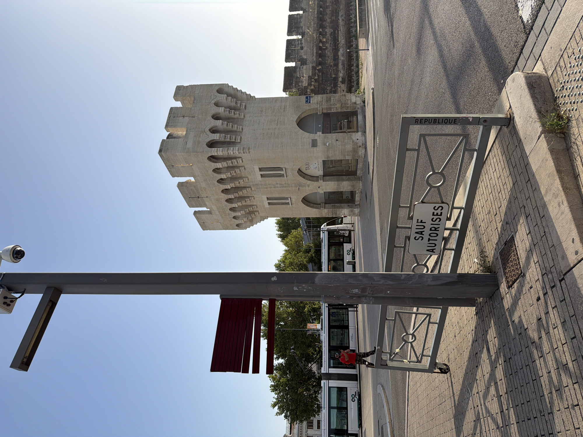 This is a street-level, medium-shot photograph taken outdoors on a clear day. The scene appears to be in the city of Avignon, France, as suggested by the architecture and signage. In the foreground, a grey metal pole stands vertically, supporting a light fixture and a security camera. A red, horizontal sign is attached to the pole. A metal barrier with a patterned design and wheels is positioned in front of the pole. To the left of the pole, a sidewalk made of brick pavers leads towards a street. A person in an orange shirt and camouflage pants is walking on the sidewalk. In the background, a tram is visible on the street. To the right of the tram and further back, a light-colored stone tower with arched openings and decorative stonework stands prominently. Adjacent to the tower is a section of a dark stone wall with battlements. The sky is clear and blue. A sign on the barrier reads "SAUF AUTORISE". Another sign on the tower reads "TAXI".
