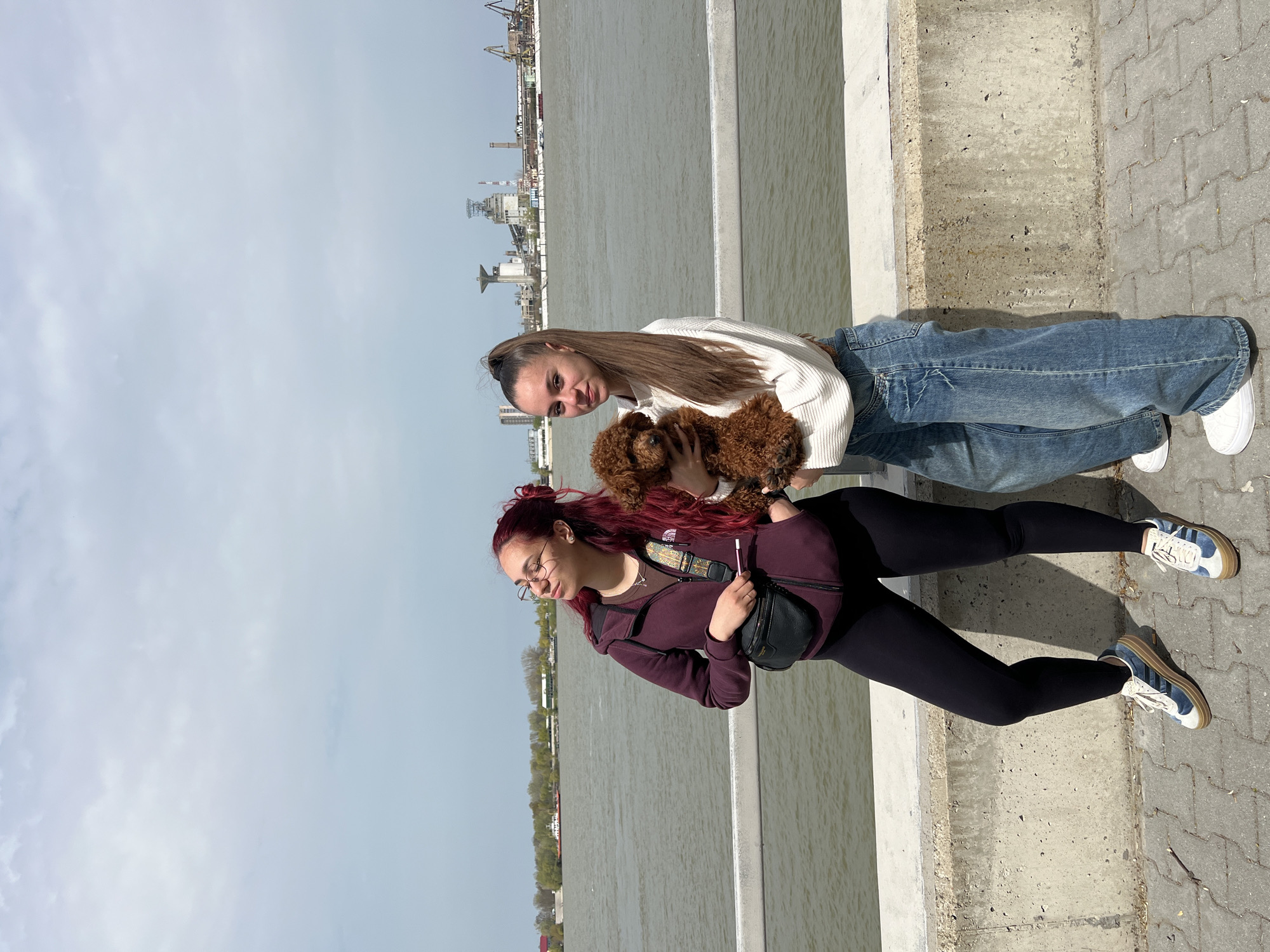 Two women are standing on a concrete ledge near a body of water in Nikopol, Bulgaria. One woman has long brown hair pulled back in a ponytail, and she is wearing a white sweater and blue jeans. She is holding a small, brown poodle dog. The other woman has long red hair and glasses. She is wearing a maroon jacket, black leggings, and sneakers with blue stripes. She has a black fanny pack strapped across her chest. The water stretches out behind them, and in the distance, there are industrial buildings and cranes. The sky is overcast. The pavement is composed of interlocking gray bricks.