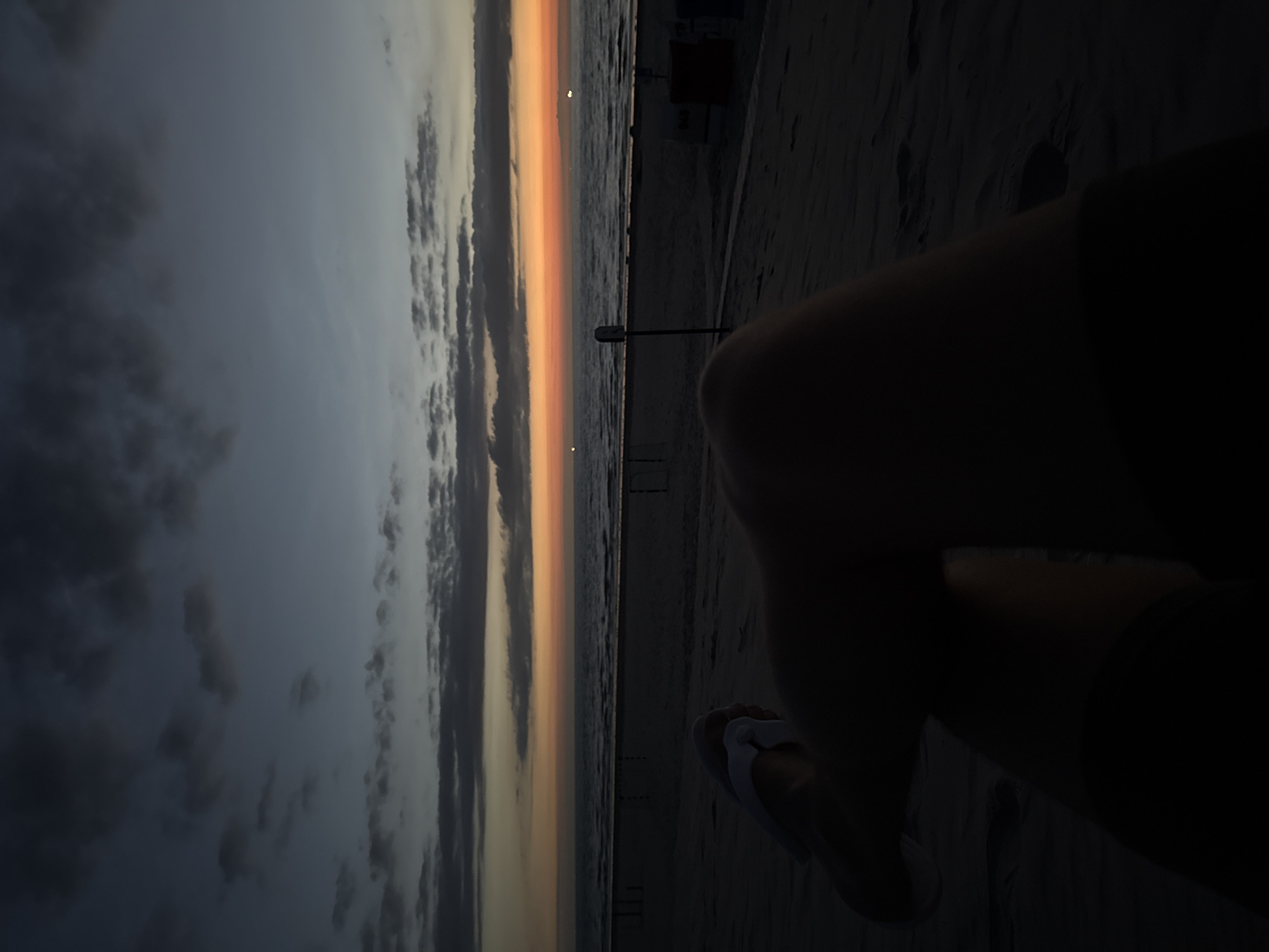 A first-person perspective shot of a person sitting on a beach at dusk. The person's legs are visible in the foreground, silhouetted against the darker sand. They are wearing white flip-flops. In the midground, the beach stretches out, with a few dark structures resembling beach chairs and a signpost. The sea is visible in the distance, with gentle waves. The horizon is marked by a band of orange and yellow light, indicative of a sunset or sunrise. Above, the sky transitions from a deep blue at the top to lighter shades of grey and blue closer to the horizon, with scattered dark clouds. The overall scene conveys a sense of peaceful solitude at the end of the day. The location is the beach on the island of Langeoog, Germany.