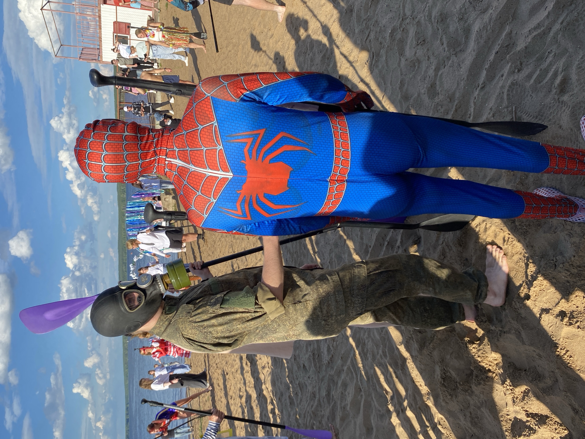 Two people are on a sandy beach near a body of water under a bright, cloudy sky. The person in the foreground is wearing a Spiderman costume and is holding a black paddle. Behind them, a person is wearing a military-style camouflage uniform and a black gas mask with goggles, also holding a paddle. The scene appears to be a public event or gathering, with other people and a small structure visible in the background. The lighting suggests it is daytime, possibly late afternoon, given the long shadows cast on the sand. The overall atmosphere seems casual and festive.
