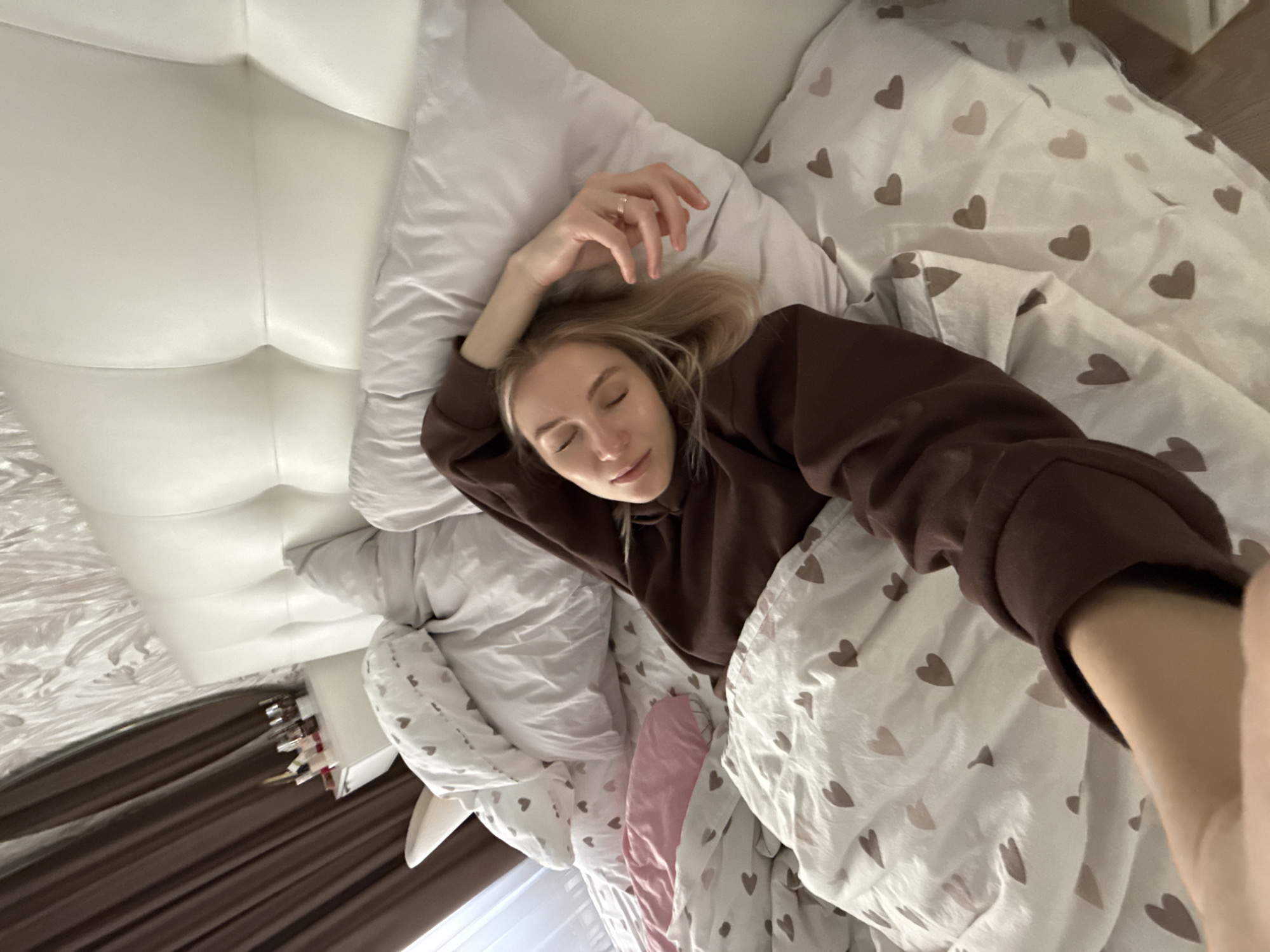 A woman lies in a bed. She has blonde hair and wears a brown sweatshirt. She has her eyes closed and one hand raised. She has a ring on her finger. The bed has white and heart patterned sheets. The headboard is white. The location is indiscernible from the image and cannot be placed within the city of Izhevsk, Russia.