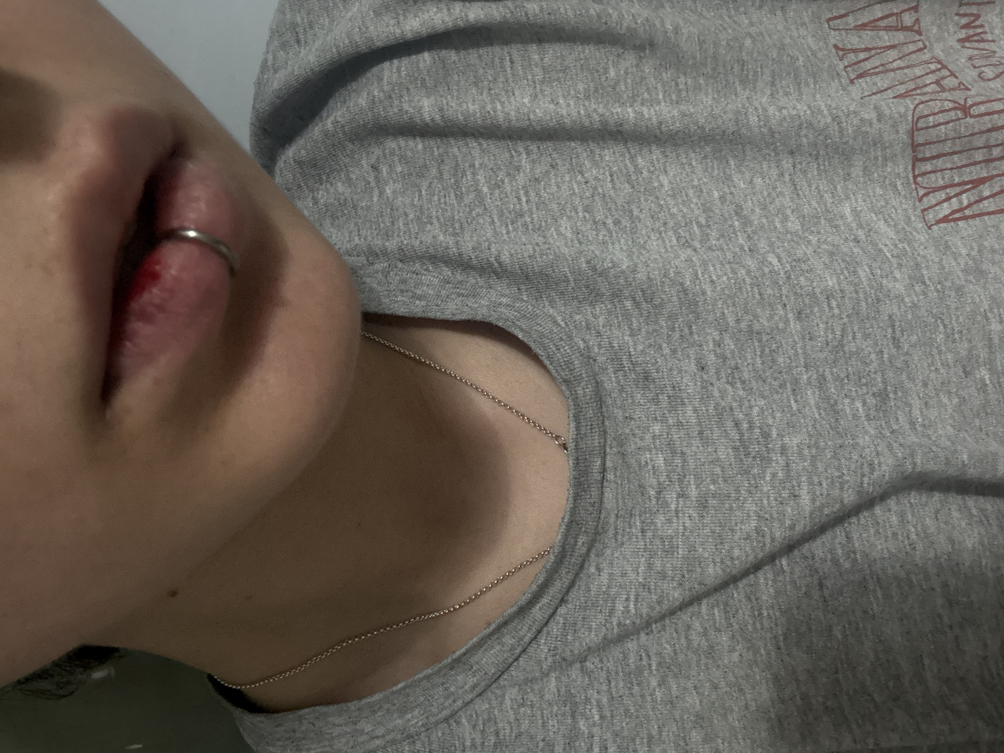The image depicts a person wearing a grey Nirvana t-shirt and a thin silver necklace. The person has a lip piercing, and there appears to be some blood or inflammation around the piercing site. The background is plain and out of focus, providing no context for a specific location in Saint Petersburg or elsewhere.