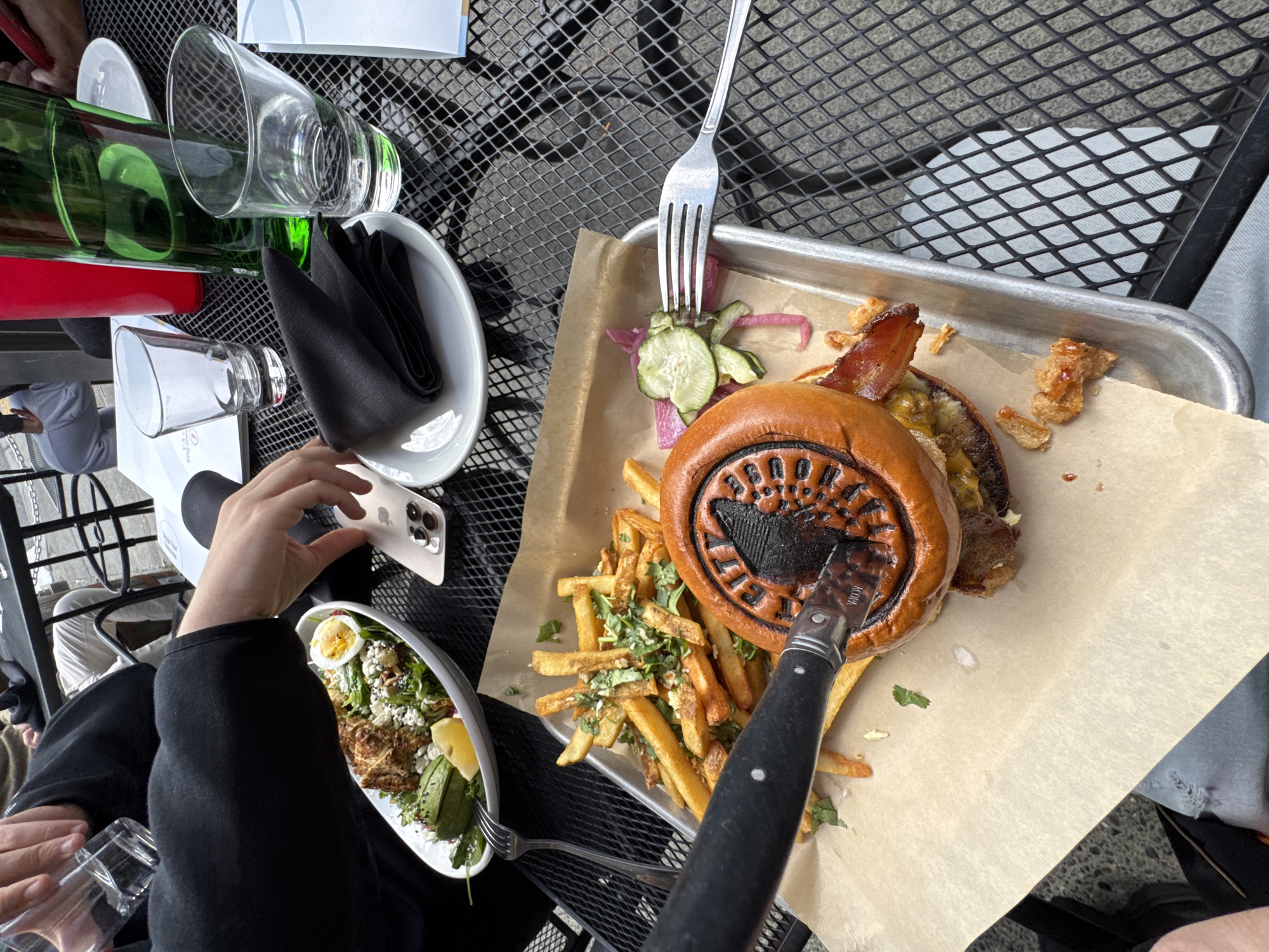 An outdoor dining scene shows a table set with a burger, fries, and a salad. A person's hand is reaching for a phone on the table. A knife is placed across the burger. In the background, a green bottle and red cup are on the table. The table is a metal mesh surface, commonly found in outdoor dining areas. This scene is likely in Anchorage, Alaska, United States of America, given the presence of a black bear motif on the burger bun.