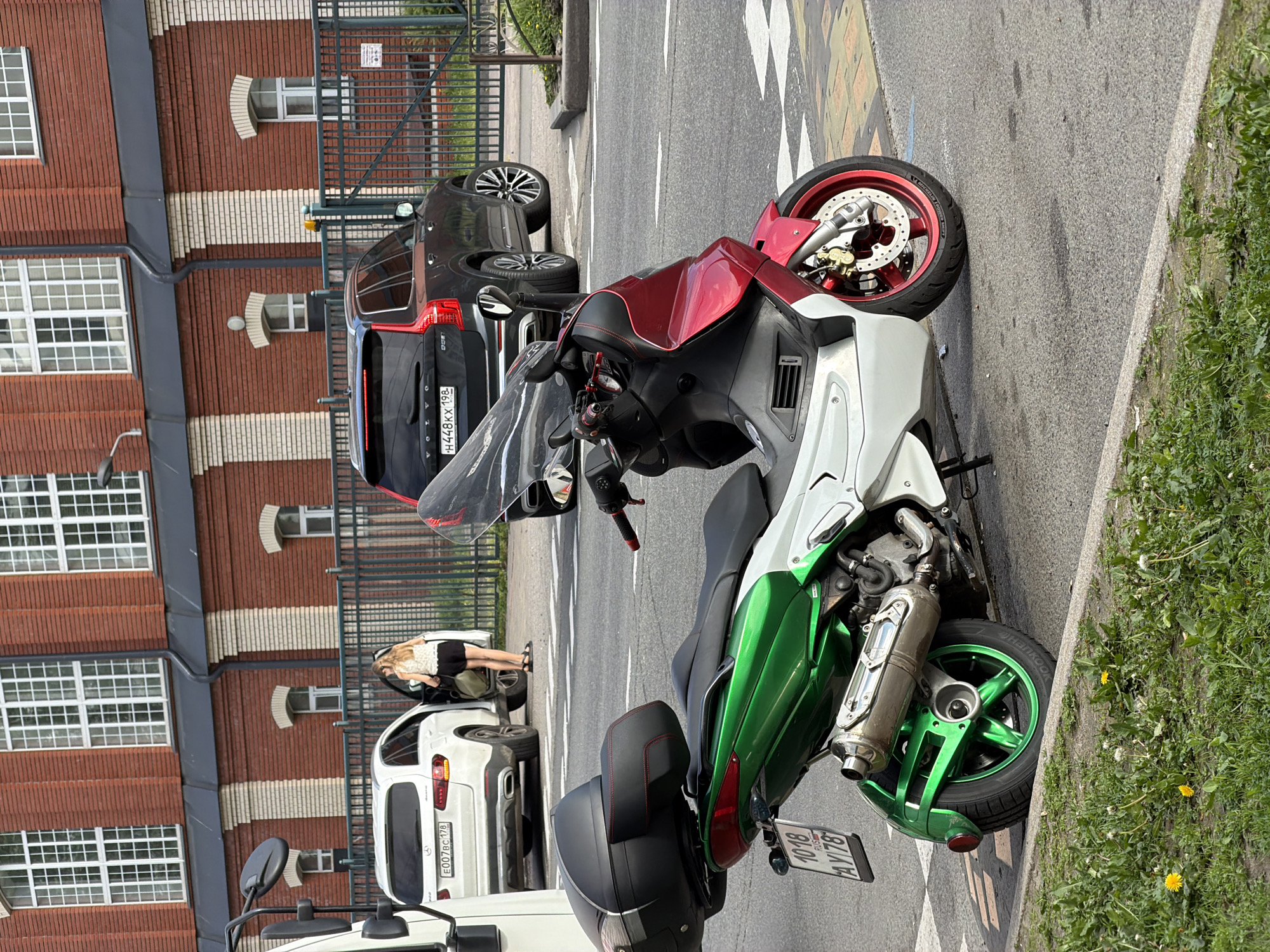 The image depicts a street scene in Saint Petersburg, Russia, with a focus on a custom-painted green and red scooter parked on the side of the road. The scooter has a black storage box on the back and a silver exhaust pipe. In the background, there is a modern brick building with large windows and a black metal fence. Several cars are also visible: a white SUV with the license plate E007BC 178, a dark grey Volvo SUV with the license plate H448KX 198, and the front of a white van on the left side of the frame. A woman is standing next to the white SUV, with her car door open. The ground is paved with asphalt, and there are yellow and white road markings. Green grass and dandelions grow along the curb.