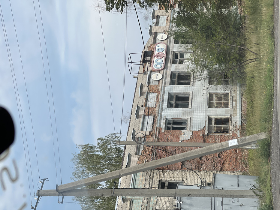 This image presents a two-story masonry building in Martove, Ukraine, showing significant damage. The left portion of the building reveals exposed red brick where the exterior cladding is absent, while the right portion retains light-colored square tiling with multiple missing sections. Numerous windows on both levels are broken or have missing panes, with the ground-floor windows on the right side secured by metal security grates. The building's upper facade features three circular signs: a central, larger sign displaying the Cyrillic word "орфе" (Orfei), flanked by two smaller signs, each bearing the Cyrillic word "ФИРМА" (FIRMA). A black, skeletal metal frame is positioned on the roof above the central sign. In the immediate foreground, a concrete utility pole with visible insulators and wires leans towards the building, partially obscuring a light-colored utility box with Cyrillic markings to the far left. The ground in front of the building is covered with overgrown grass and sparse small trees. The sky above is light blue with scattered white clouds. No people, active processes, or interactions are visible within the frame.