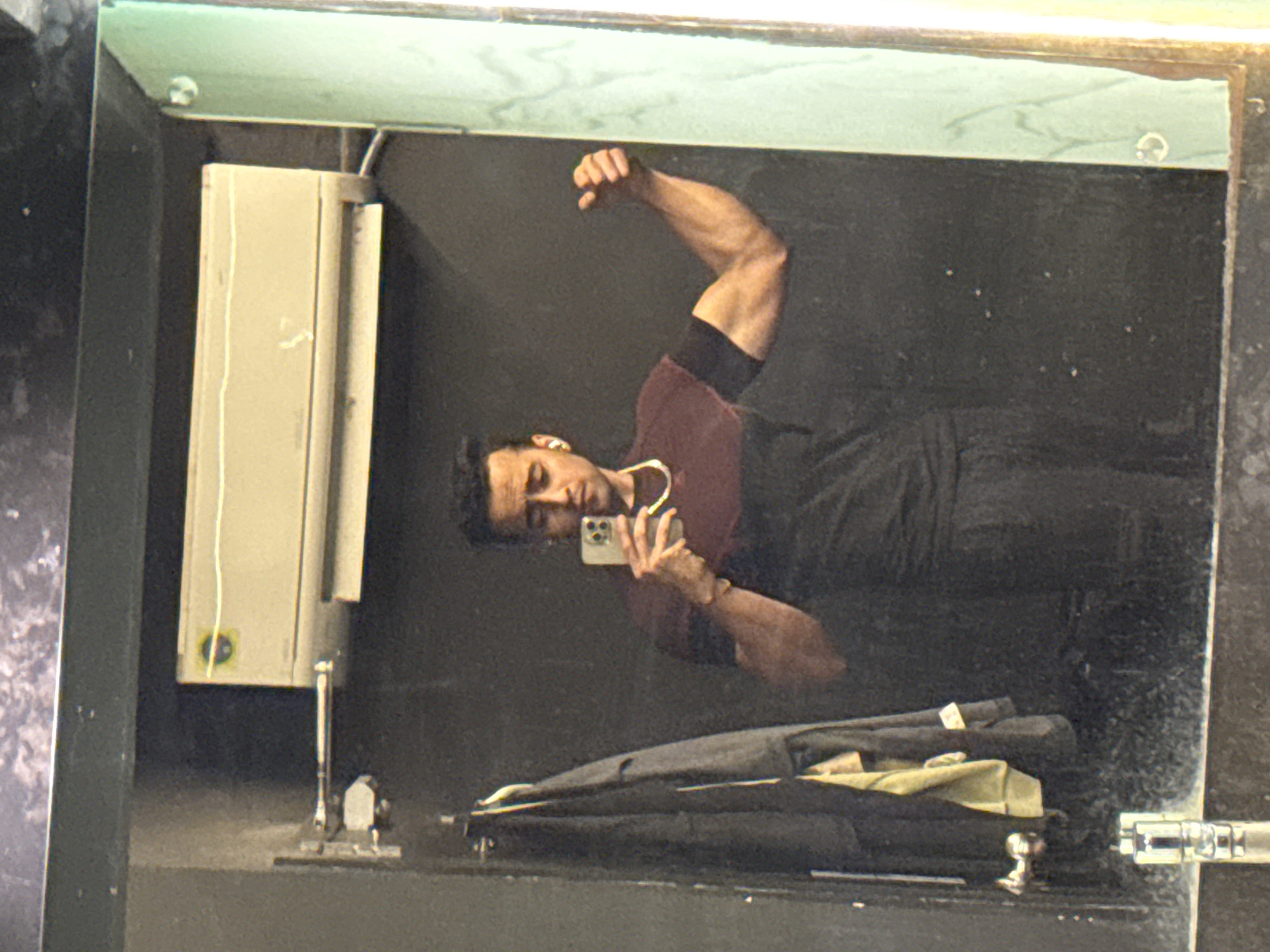 A man is taking a selfie in a bathroom mirror. He is wearing a dark shirt with burgundy sleeves and dark pants. He is flexing his right bicep. In the lower part of the image, below the man's reflection, is a dark garment bag and a light-colored cloth. To the left of the mirror, mounted on the wall, is a light-colored dispenser. The location of the image is not provided in the context or visual information. Therefore, determining the location in Kolkata, India is not possible.