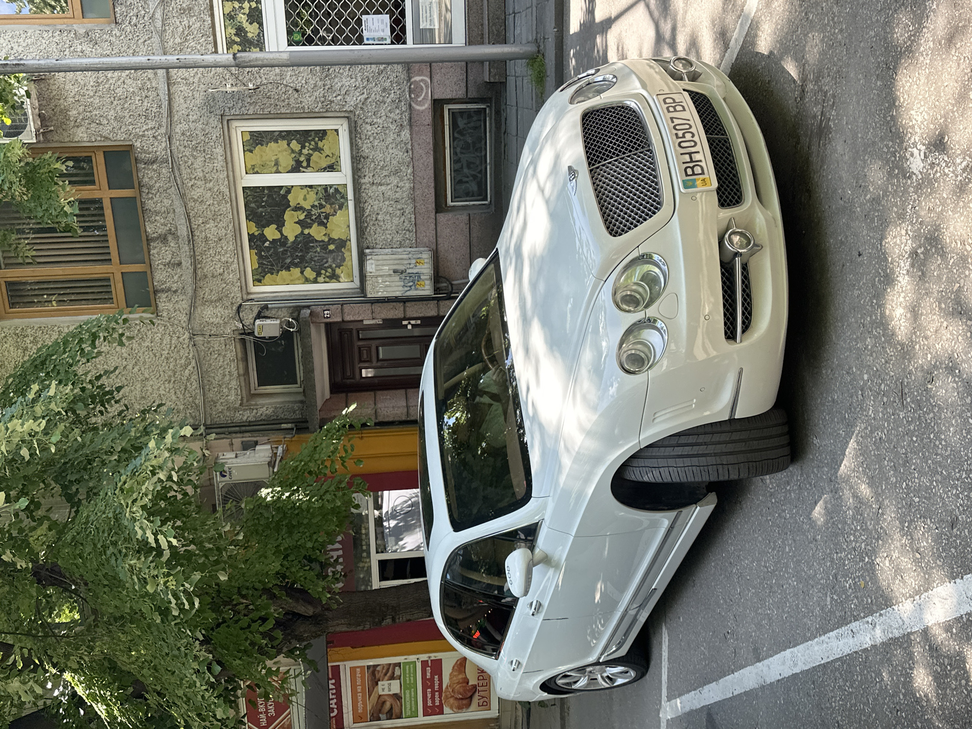 The image shows a white Bentley parked on a street with a white line. The license plate reads "BH 0507 BP" and shows the Ukrainian flag. Behind the car is a building with several windows and a door, and a tree with green leaves partially obscures the building. A sign for "CARA" is visible, advertising baked goods, and listing them as "healthy". The sign is written in Ukrainian. The location appears to be in Ukraine.