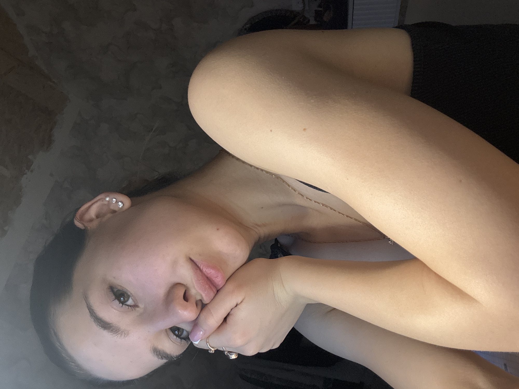 The image shows a young woman with fair skin, dark hair, and brown eyes. She is wearing a black top and a gold necklace. Her left hand is positioned near her mouth, adorned with two rings. She has multiple piercings in her left ear. The background is a dark, textured surface. The lighting is uneven, with a bright light source highlighting her skin. The image does not show any recognisable elements of Noviy, Russia, so it cannot be identified as the location.