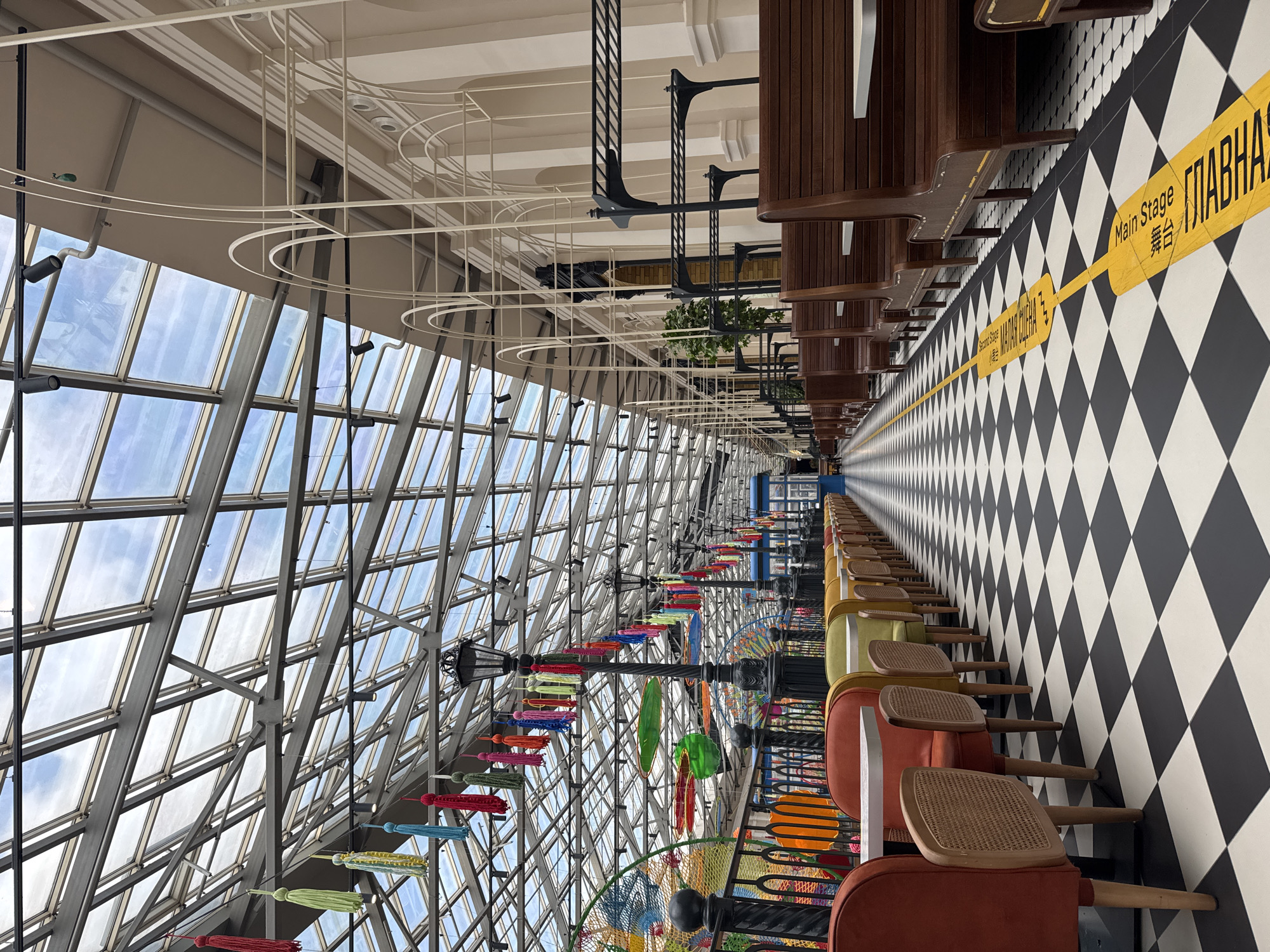The image depicts the interior of a brightly lit, spacious establishment with a retractable glass ceiling. The floor is a striking black and white checkered pattern, and a yellow line with text runs down the center. To the left, a row of colorful chairs with wooden legs and rattan seats are arranged around tables. These chairs are upholstered in shades of red, green, and yellow. Beyond them, a railing with decorative fencing encloses an area with colorful, playful decorations, including a net-like structure and what appear to be surfboards. Several black lampposts adorned with colorful tassels are spaced along this railing.
To the right, a long row of dark wooden booth-style seating with white tabletops stretches into the distance. The walls are a light cream color, featuring a series of arched decorative elements. Above, the glass ceiling reveals a cloudy sky, supported by a metal framework. The overall ambiance is cheerful and inviting, suggesting a public space like a restaurant, cafe, or entertainment venue. No people are visible in the scene.