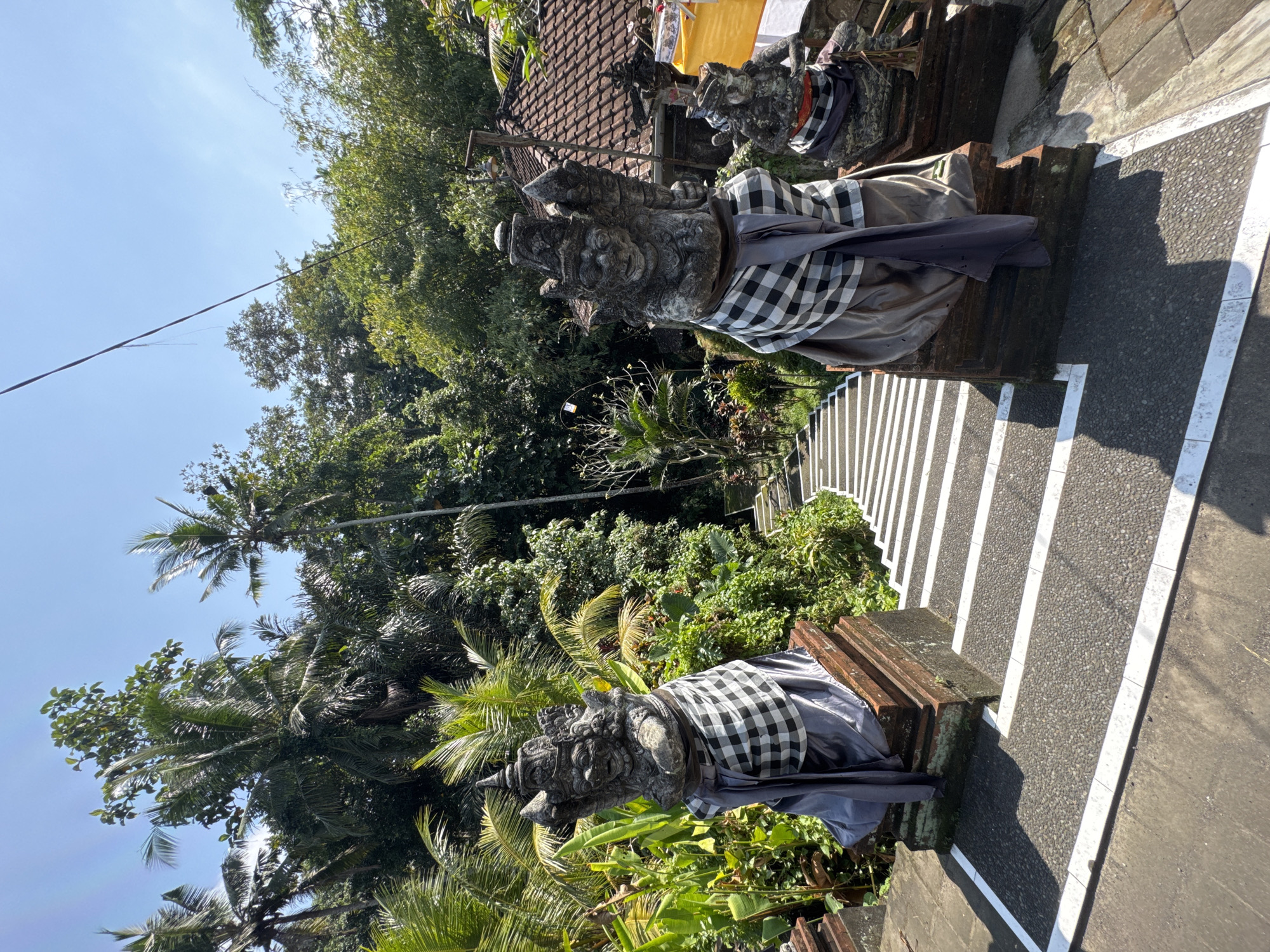 This image depicts a set of stone statues at the entrance to a staircase in Ubud District, Indonesia. The statues, carved in a traditional Balinese style, are adorned with checkered fabric wraps. They are positioned on pedestals flanking the top of the stairs. The staircase itself is made of stone with white markings on the edges of each step and is surrounded by lush, dense greenery, including palm trees. The sky above is clear and blue. The overall scene suggests a cultural or religious site in a tropical environment.