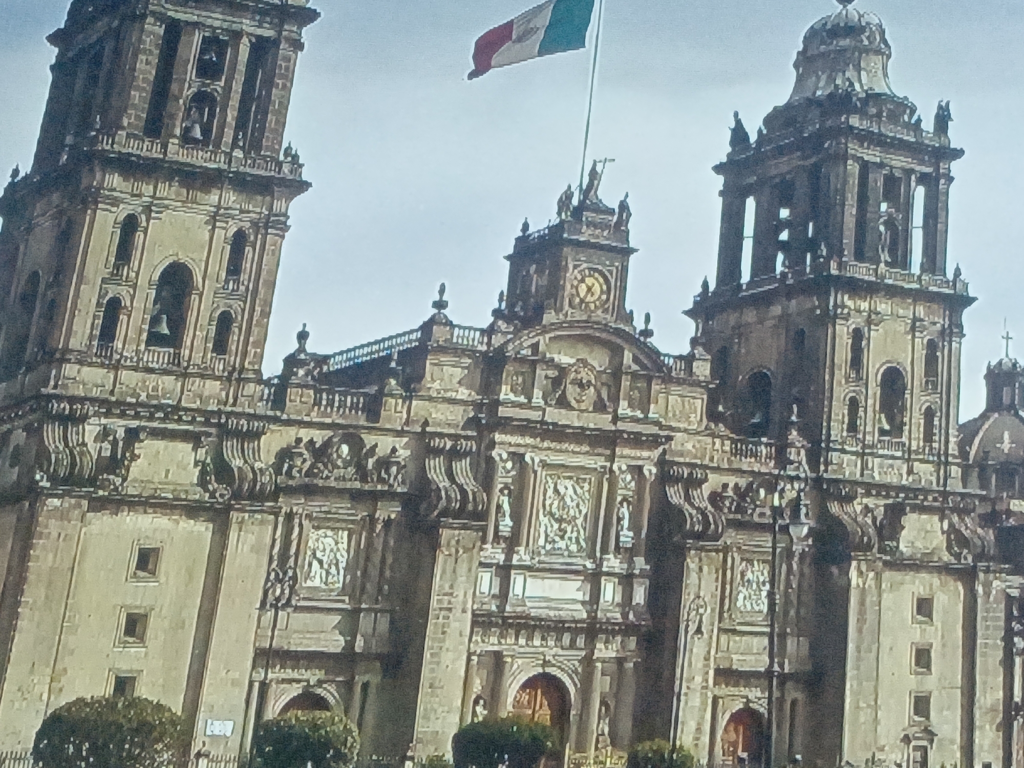 The image shows the Metropolitan Cathedral of the Assumption of Mary of Mexico City, a large, ornate cathedral featuring two bell towers and a central clock tower. A Mexican flag flies above the cathedral. The building is constructed of light-colored stone. In the foreground, some trees are present. The sky is overcast. The image does not appear to be located in Juan Griego, Venezuela.