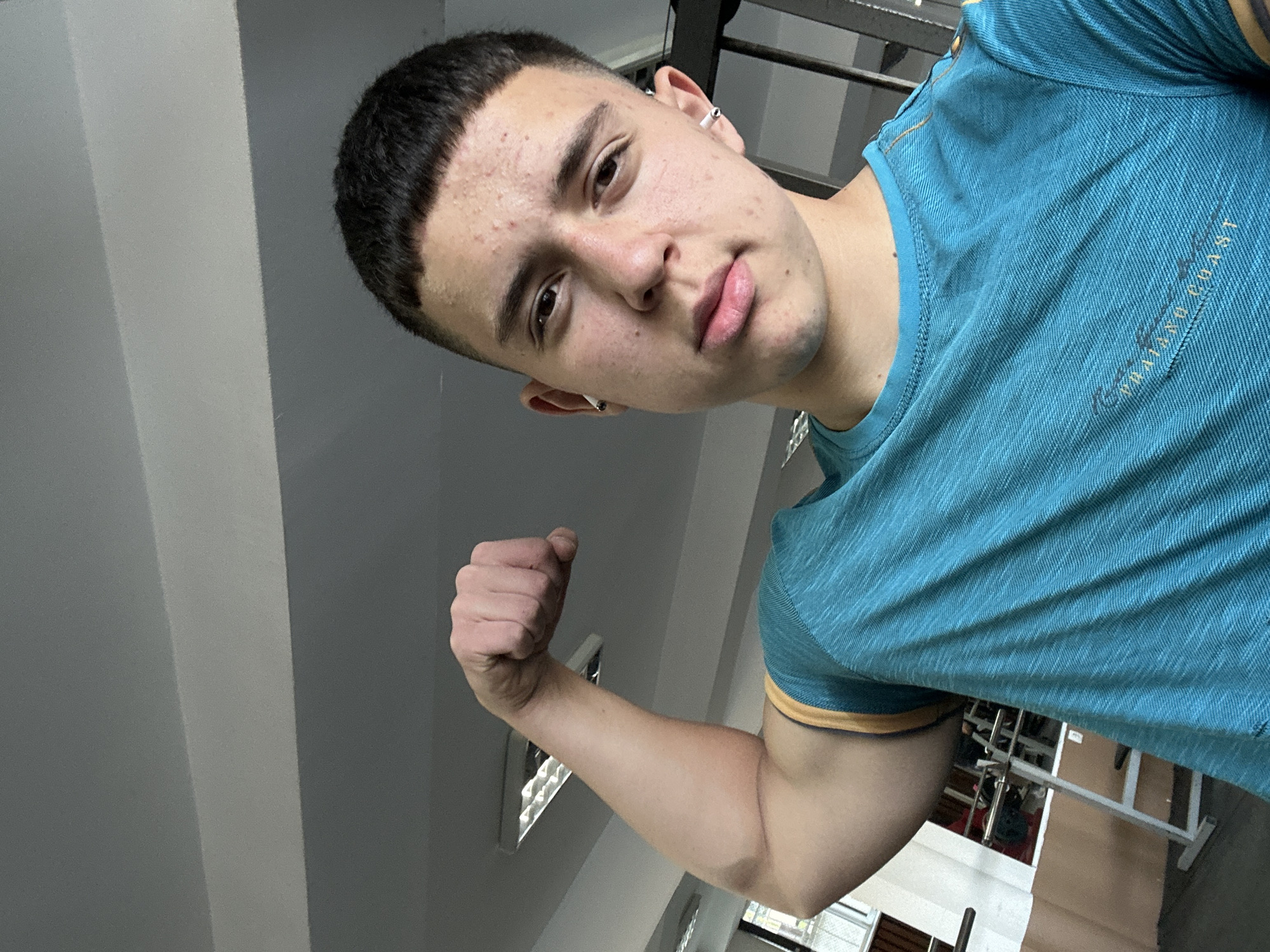 An angled, medium-shot portrait depicts a young male indoors, possibly in a gym in Zlatograd, Bulgaria. He looks directly at the camera with a slightly serious or pouting expression. He has short, dark hair with shaved sides, and his face shows some acne. He wears small white in-ear headphones or earbuds and has ear piercings.
He is dressed in a blue t-shirt with a textured, striped pattern and a gold or dark yellow trim on the collar and sleeves. Faint text reading "VALLEJO COAST" is visible on the right side of his chest. The subject is flexing his right bicep, holding a closed fist.
The background features light grey walls and a ceiling with white rectangular light fixtures. Distant metallic structures, resembling gym equipment, are partially visible, suggesting a fitness facility. The lighting is consistent with an indoor environment.