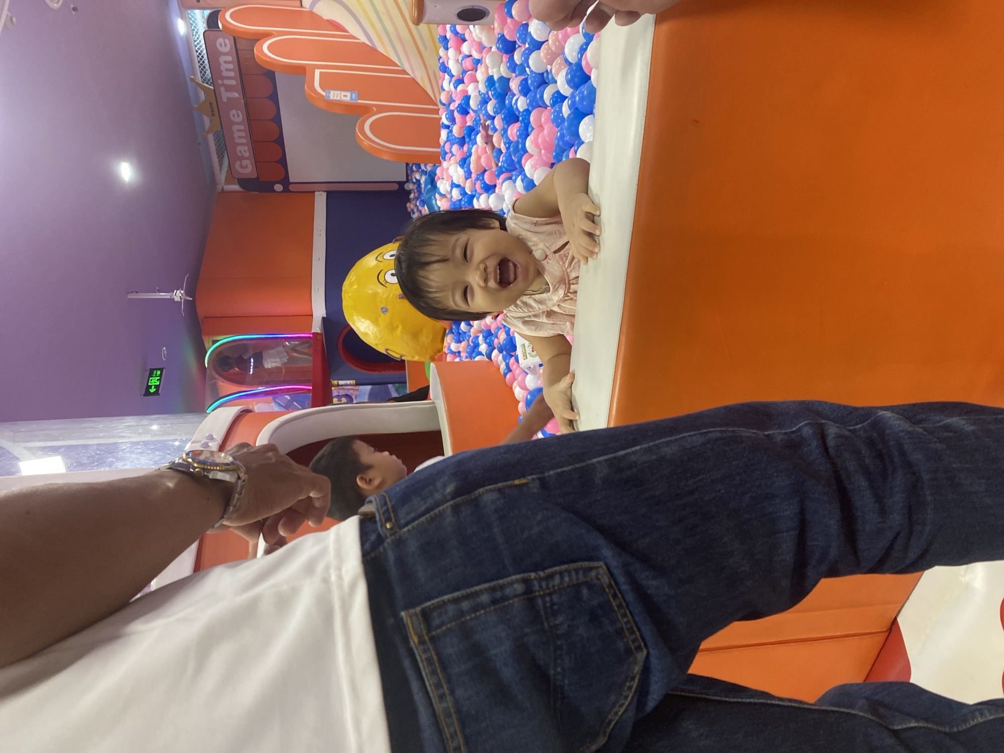 The image shows a toddler with dark hair, wearing a light-colored sleeveless top, laughing joyfully while peeking over the edge of an orange barrier in a ball pit. The ball pit contains numerous blue, pink, and white plastic balls. Behind the child, a large yellow inflatable object with a face is visible. Further in the background, a sign reads "Game Time" with an orange and white striped border. The scene appears to be an indoor children's play area. A person's arm and shoulder, wearing a white t-shirt and denim jeans with a watch on the wrist, are visible in the foreground, suggesting they are interacting with or observing the child. The location is identified as Phu Thuy, Vietnam.