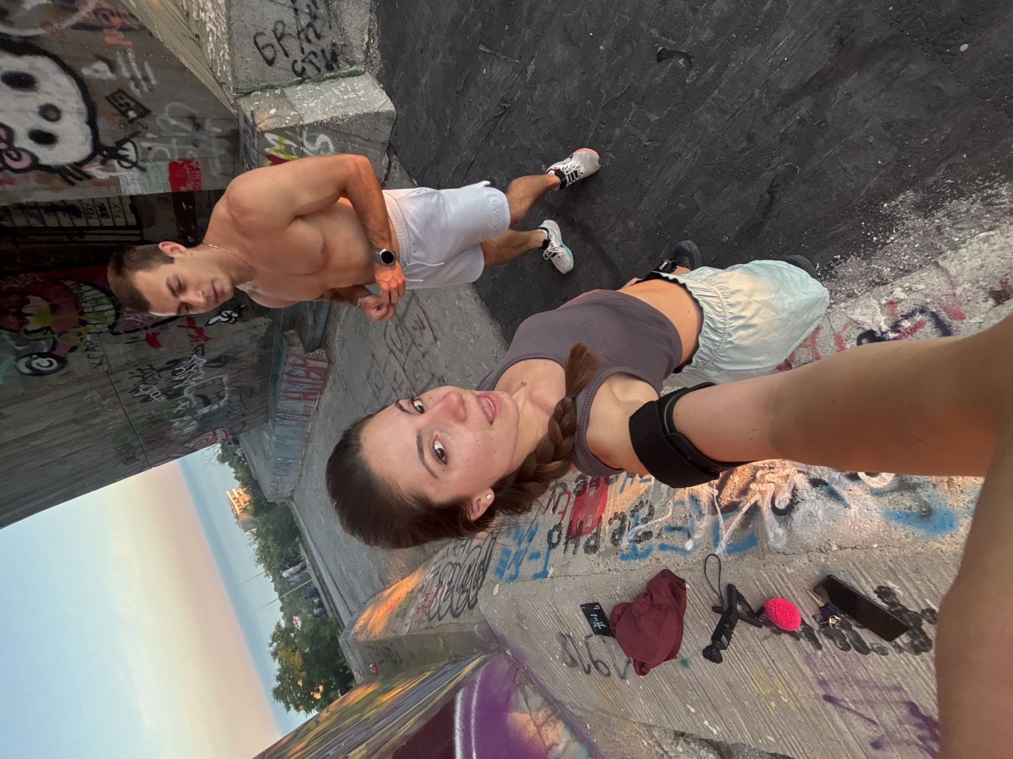 A high-angle, wide selfie captures a young Caucasian couple standing on a concrete structure at sunset. The woman, in the foreground, is wearing a gray crop top and light blue shorts. She has a braid down her back and is holding the camera out. The man, behind her, is shirtless and wearing white shorts. Both are wearing athletic shoes. They are on a concrete platform covered in graffiti, with a view of the Black Sea in the distance. The sunset casts a warm glow on the scene. The location is confirmed to be Varna, Bulgaria, specifically on the "Mali" concrete structure.