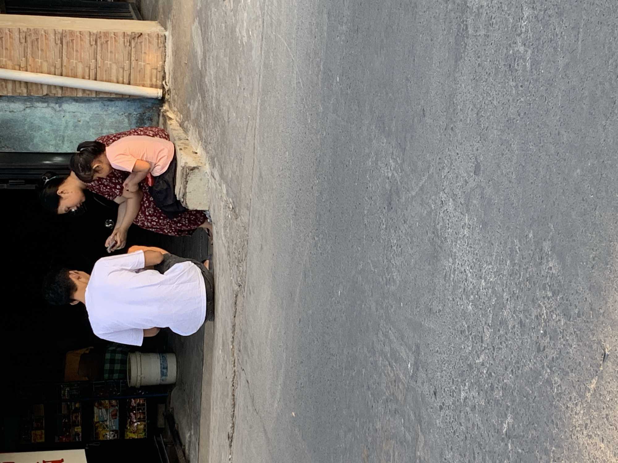 This is a full-body shot of three individuals in what appears to be a street scene in Makassar, Indonesia. In the foreground, a man wearing a white t-shirt and dark pants is crouched down, facing away from the camera, with his hands engaged with something on the ground. To his right, a woman in a patterned dress is seated on a concrete ledge, holding a mobile phone. Next to her, a young girl in an orange t-shirt and dark pants is also seated, looking down at her hands. Behind them is a dark storefront with shelves stocked with various goods. The ground is paved with asphalt.