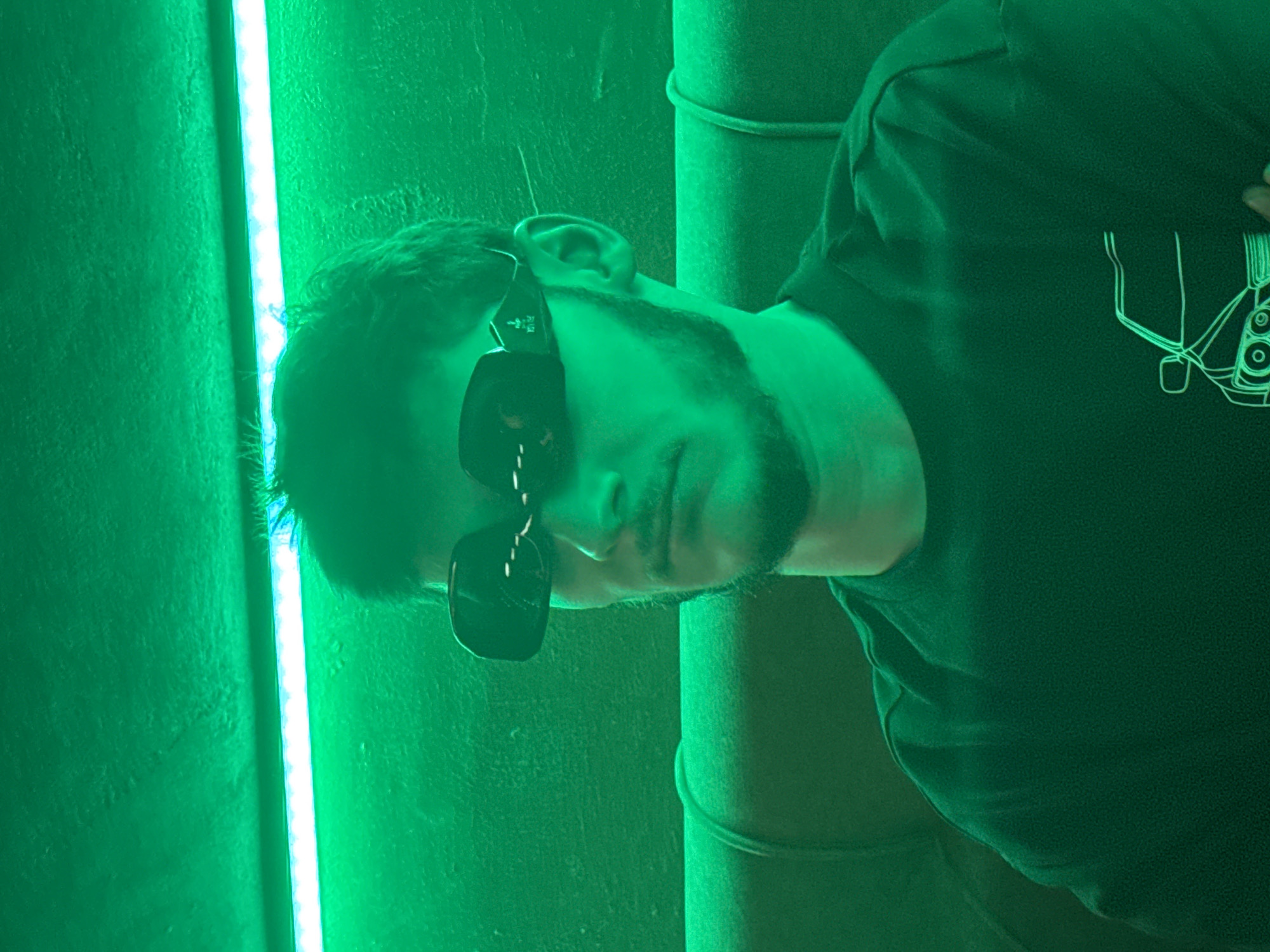 A man with a beard and dark hair is posing in front of a green light source. He is wearing dark sunglasses and a dark t-shirt with a white design on the right side. He is standing next to what appears to be a vertical metal pipe. The scene is lit with a strong green light. It is difficult to determine the location in Sofia, Bulgaria based on the information provided.