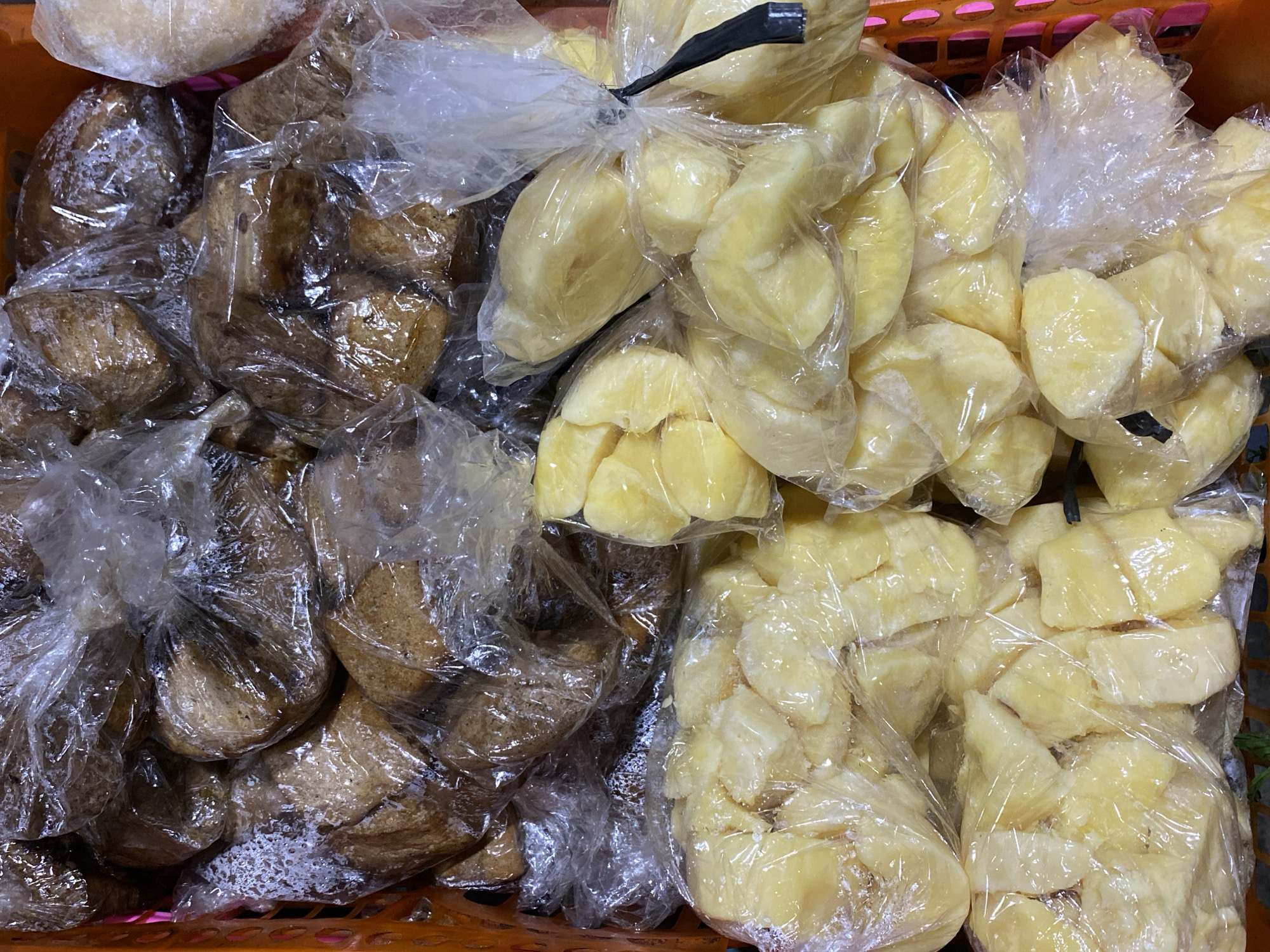 An overhead, close-up shot displays a collection of food items in plastic bags, arranged within an orange plastic crate. The crate is filled with baked goods on the top half, appearing to be small, round loaves of bread, some of which show signs of mold. The bottom half of the crate is filled with chopped pieces of what appears to be durian fruit, each portion contained within its own plastic bag. The durian pieces are a pale yellow color and appear to be fresh. The lighting is even, and there is no indication of people or activity in the scene. The image provides no information about the location within Ambarketawang, Indonesia, or any specific details about the food's origin or purpose.