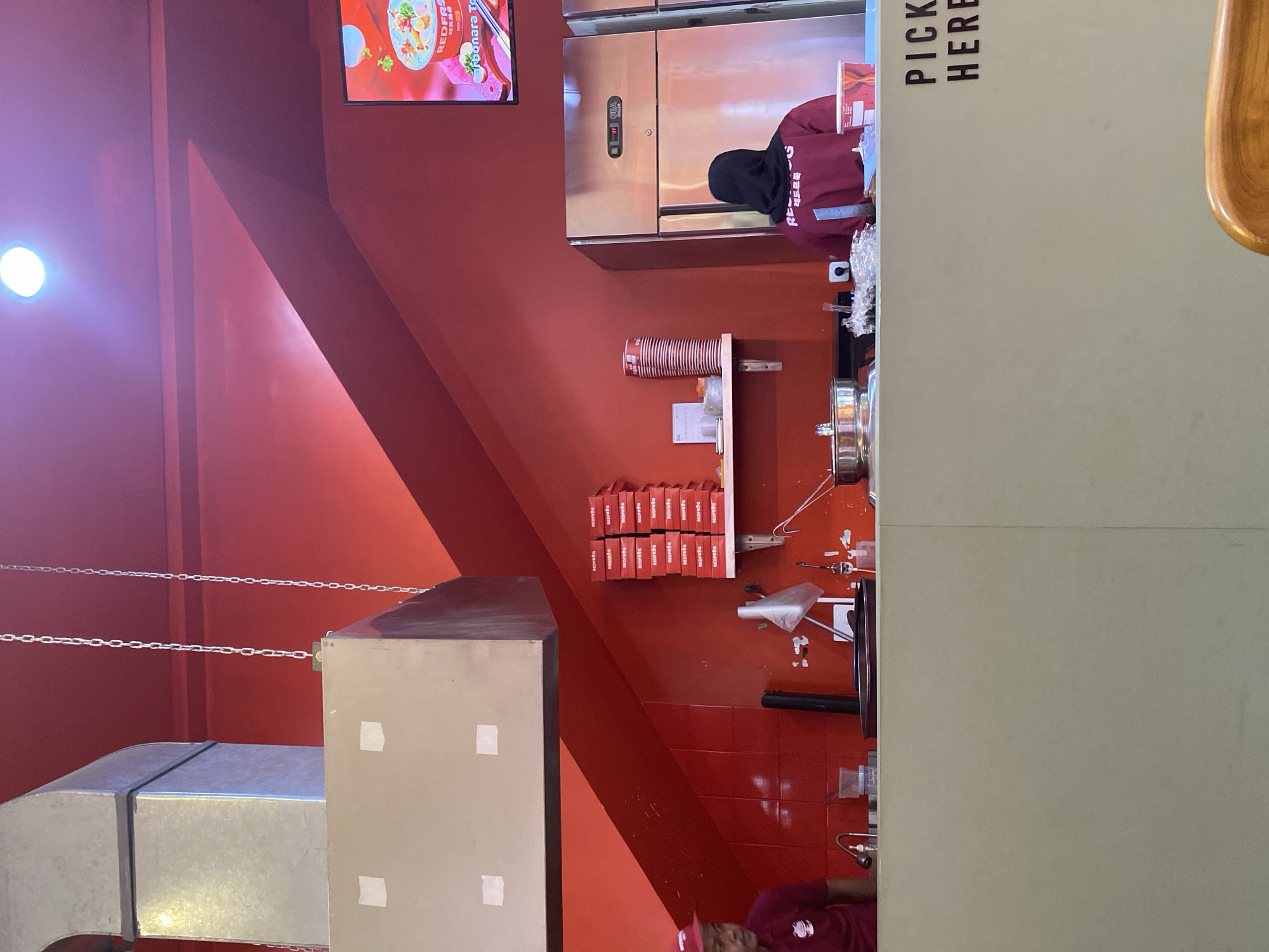 The image depicts the interior of a food establishment in East Jakarta, Indonesia, likely a quick-service restaurant given the setup. The dominant color scheme is red, with red walls and a red ceiling that features a bright light fixture. A large metallic ventilation hood hangs from the ceiling. In the foreground, a counter separates the service area from the customer space. On the left, a person wearing a maroon uniform and a red cap is visible behind the counter, near a sink area. On the right, a person wearing a maroon hijab and a maroon top with text on the back is standing behind the counter, near a large stainless steel refrigerator. Stacked red boxes with white lettering are neatly arranged on a shelf on the right wall. A television screen displaying food imagery is mounted high on the right wall. The counter surface is cluttered with various items, including a pot, plastic wrap, cups, and what appears to be a small calendar. The words "PICK HERE" are printed in black capital letters on the light-colored counter in the foreground.