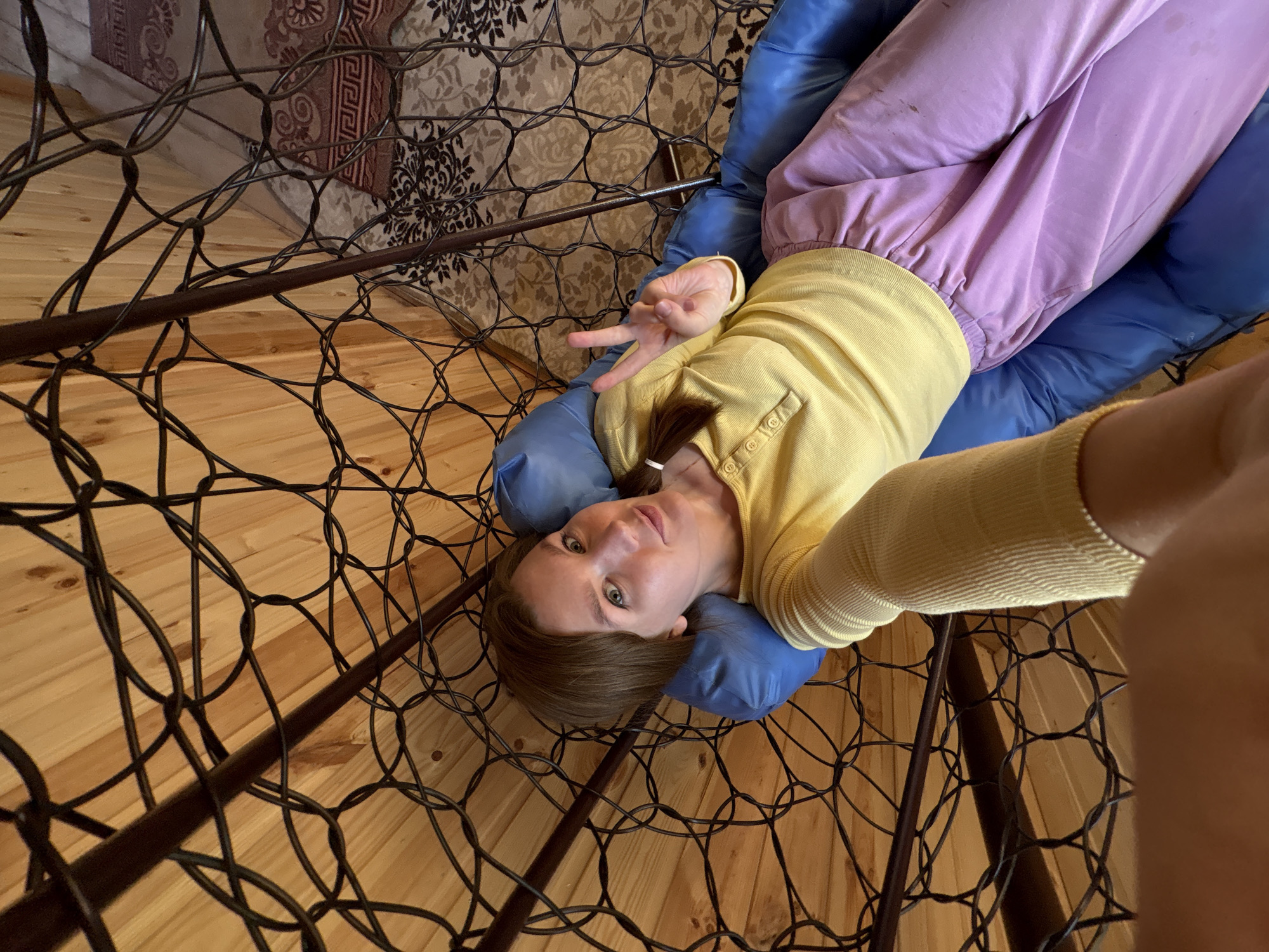 A woman is lying on a blue cushion within a metal-framed structure. She is wearing a yellow long-sleeved shirt and lilac sweatpants. She is making a peace sign with her fingers. The location is Italmas, Russia, as per metadata. The background includes wooden flooring and a wall. The image appears to be taken from a slightly elevated perspective.
