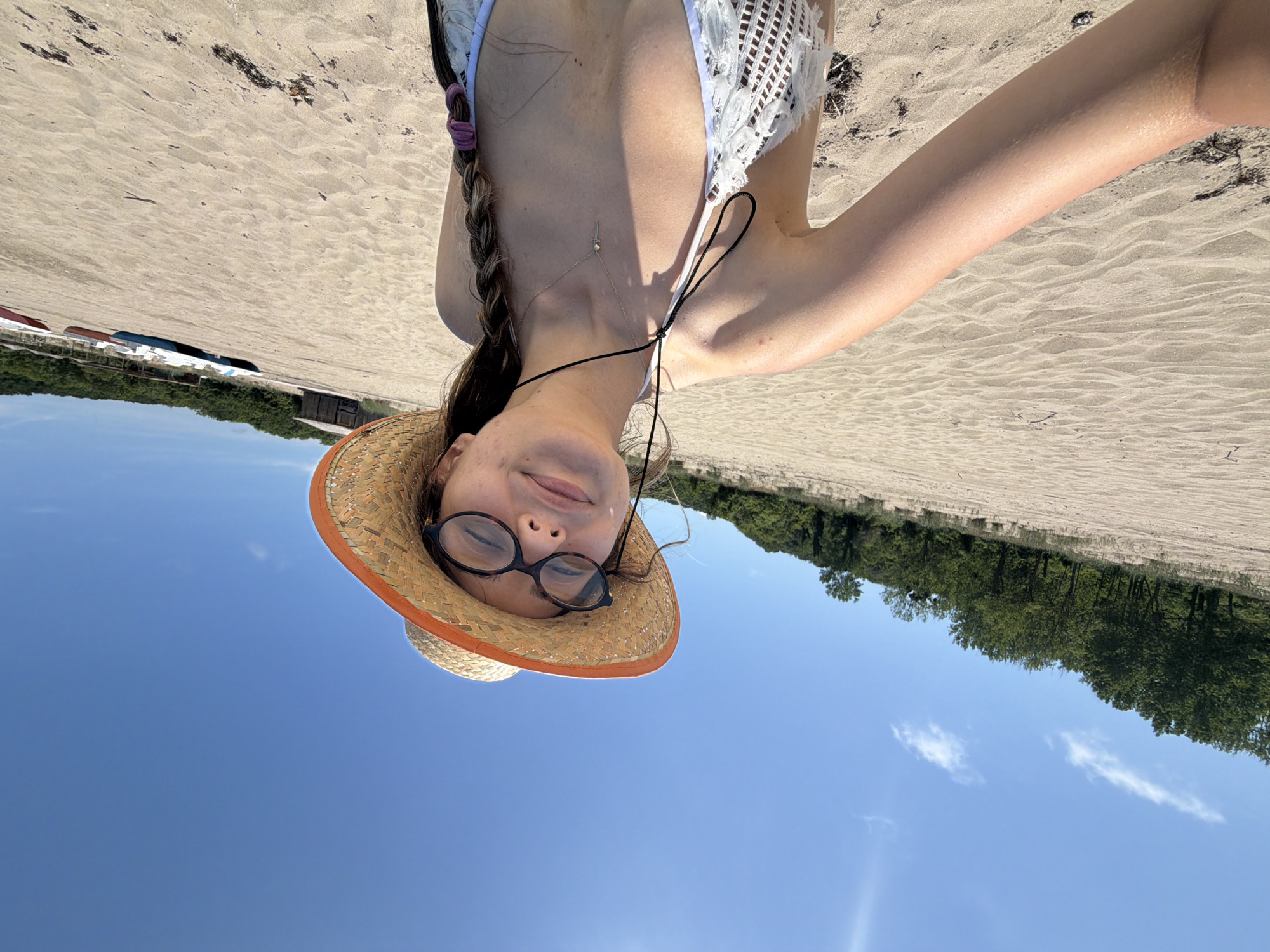 A woman with long dark hair braided in a plait, wearing glasses and a straw hat with an orange band, is taking a selfie on a beach. She is wearing a white bikini top. The beach sand is visible, with some seaweed and debris. In the background, there is a line of trees and a clear blue sky with a few wispy clouds. Several small boats are lined up on the sand in the distance. The location is the beach in Rezovo, Bulgaria.